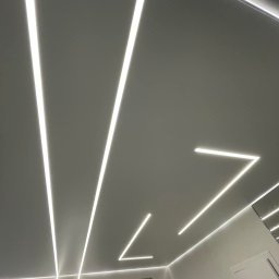 MASTERBUD MAKSYM ROZUMNIAK - Nowoczesne oświetlenie sufitowe LED z liniowymi lampami tworzącymi geometryczne wzory w białym wnętrzu, widoczne okno i fragment lustra.