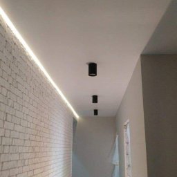 MASTERBUD MAKSYM ROZUMNIAK - Korytarz z oświetleniem LED wzdłuż ściany z cegły i trzema czarnymi reflektorami sufitowymi.
