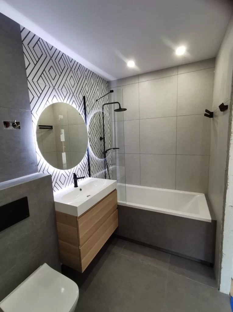 Nowoczesna łazienka z geometryczną tapetą, okrągłym lustrem z podświetleniem LED, drewnianą szafką pod umywalką i czarną armaturą.