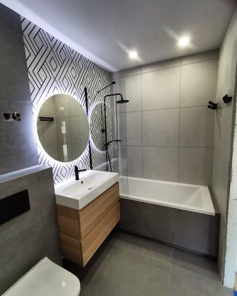 Nowoczesna łazienka z okrągłym lustrem podświetlanym LED, białą umywalką na drewnianej szafce, szarą glazurą, czarną armaturą i geometryczną tapetą w tle.