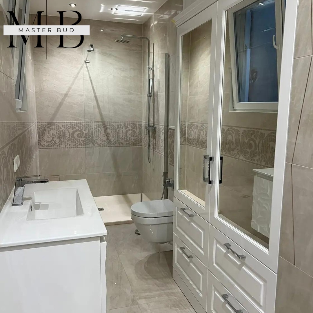 Jasna łazienka z beżowymi płytkami, kabiną prysznicową typu walk-in, umywalką z szafką i wysoką szafą z szufladami oraz oknem uchylnym.