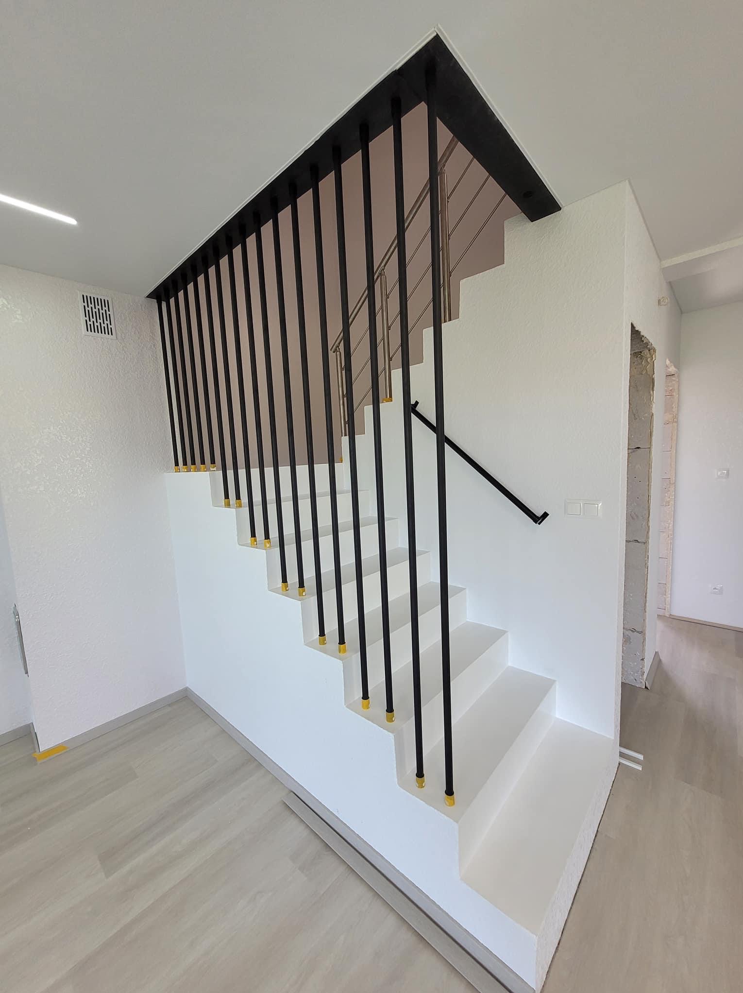 Nowoczesne, białe schody z czarną balustradą i złotymi detalami, wykończone w jasnym wnętrzu z teksturowanymi ścianami i drewnopodobną podłogą.