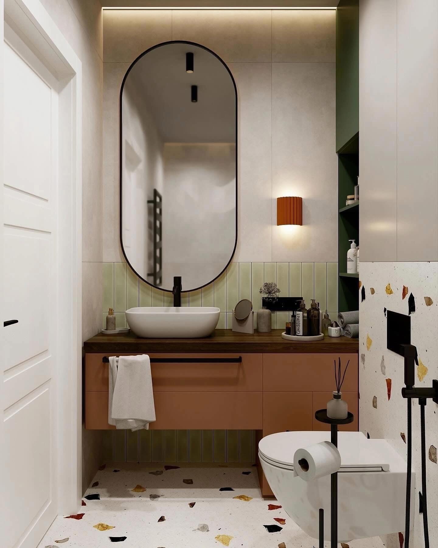 Nowoczesna łazienka z owalnym lustrem w czarnej ramie, białą umywalką nablatową, szafką w kolorze brzoskwini, toaletą z czarnym uchwytem na papier i podłogą w stylu terrazzo.