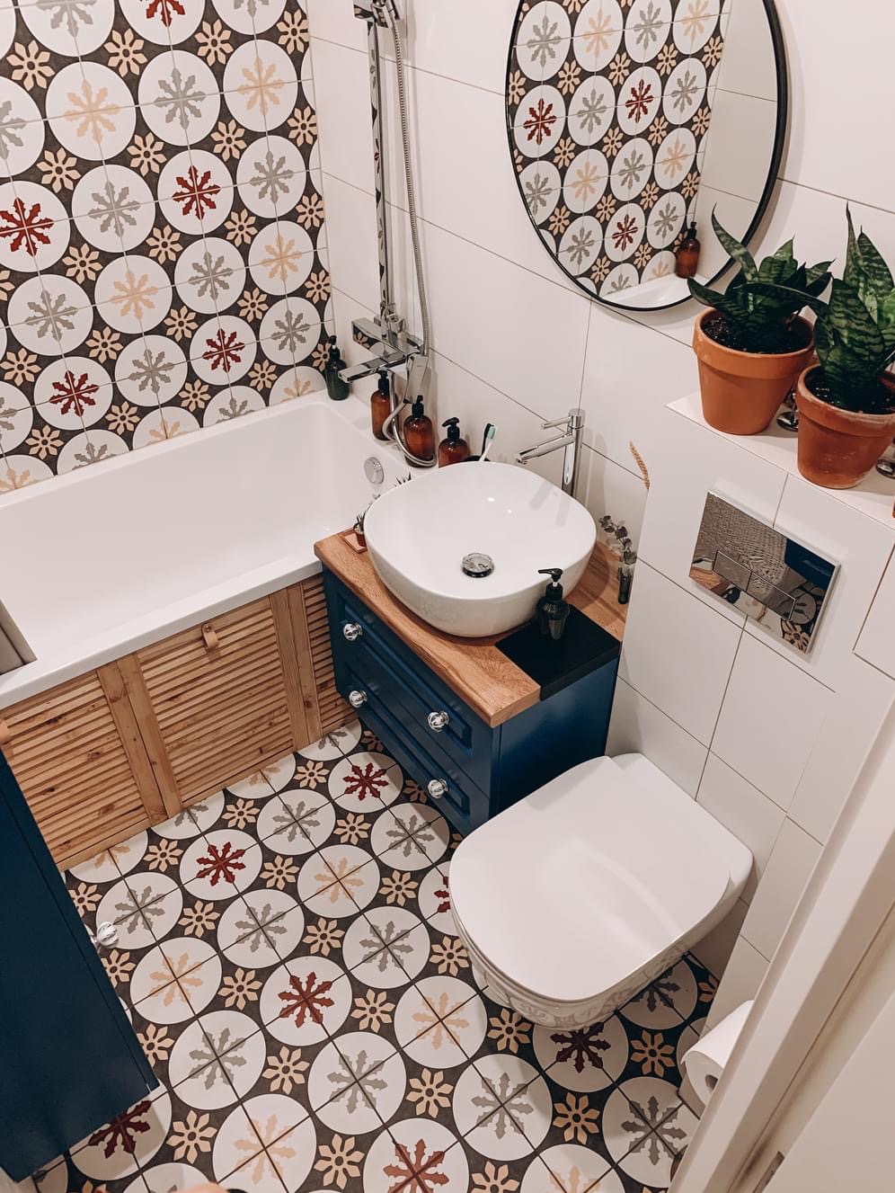 Wnętrze wykończonej łazienki w Warszawie z wanną obudowaną drewnianymi lamelami, umywalką nablatową na niebieskiej szafce z drewnianym blatem, toaletą oraz wzorzystymi płytkami podłogowymi...