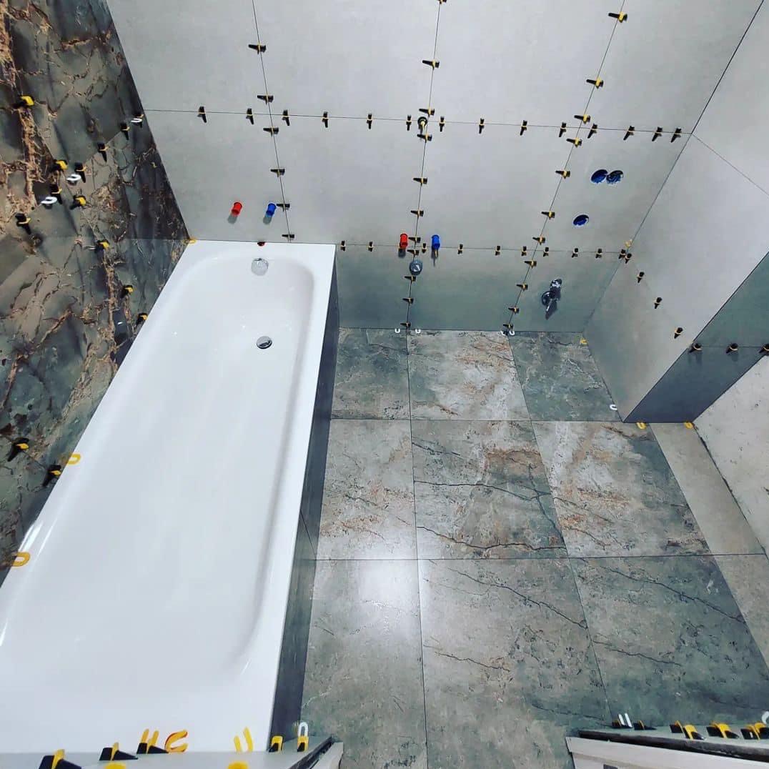 Układanie płytek w łazience z użyciem systemu poziomowania, widoczna wanna i przygotowane otwory na armaturę.