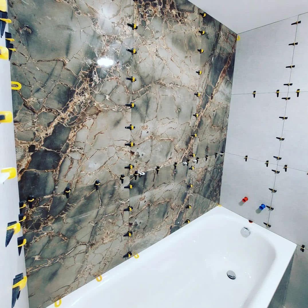 Układanie płytek imitujących marmur w łazience, widoczne klipsy poziomujące i krzyżyki dystansowe, biała wanna w rogu.