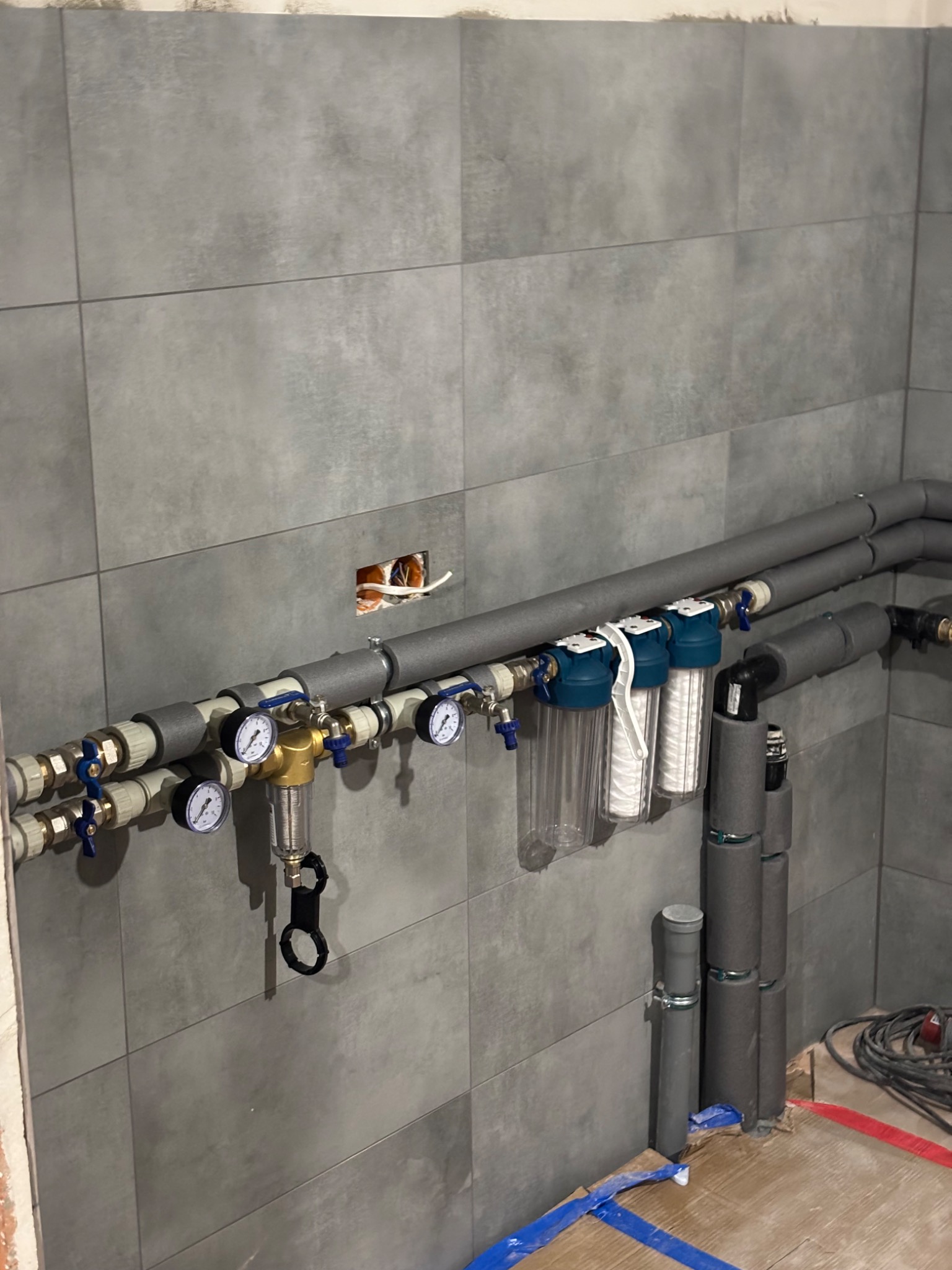 Szare płytki na ścianie w pomieszczeniu w trakcie remontu. Widoczne rury instalacji wodnej z manometrami, zaworami, filtrami wody i izolacją termiczną. Część instalacji jest jeszcze niepodłączona...
