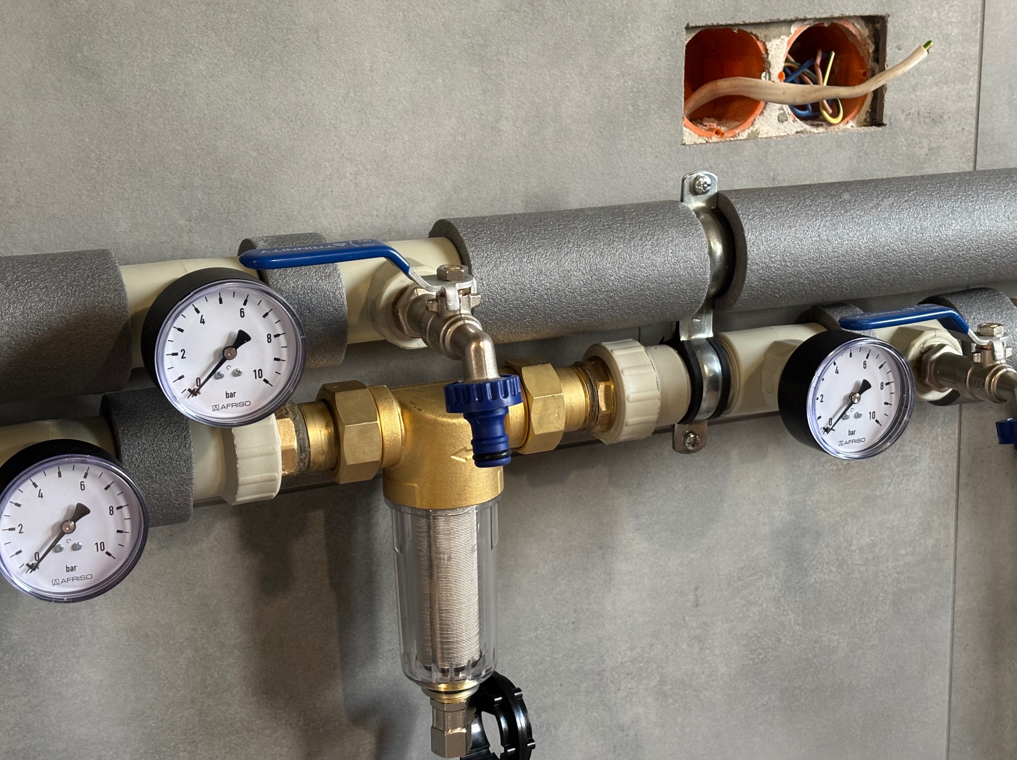 Instalacja hydrauliczna z manometrami i filtrem, zabezpieczona izolacją termiczną, widoczna na tle szarej ściany w Piekary. W ścianie otwór z wystającymi przewodami.