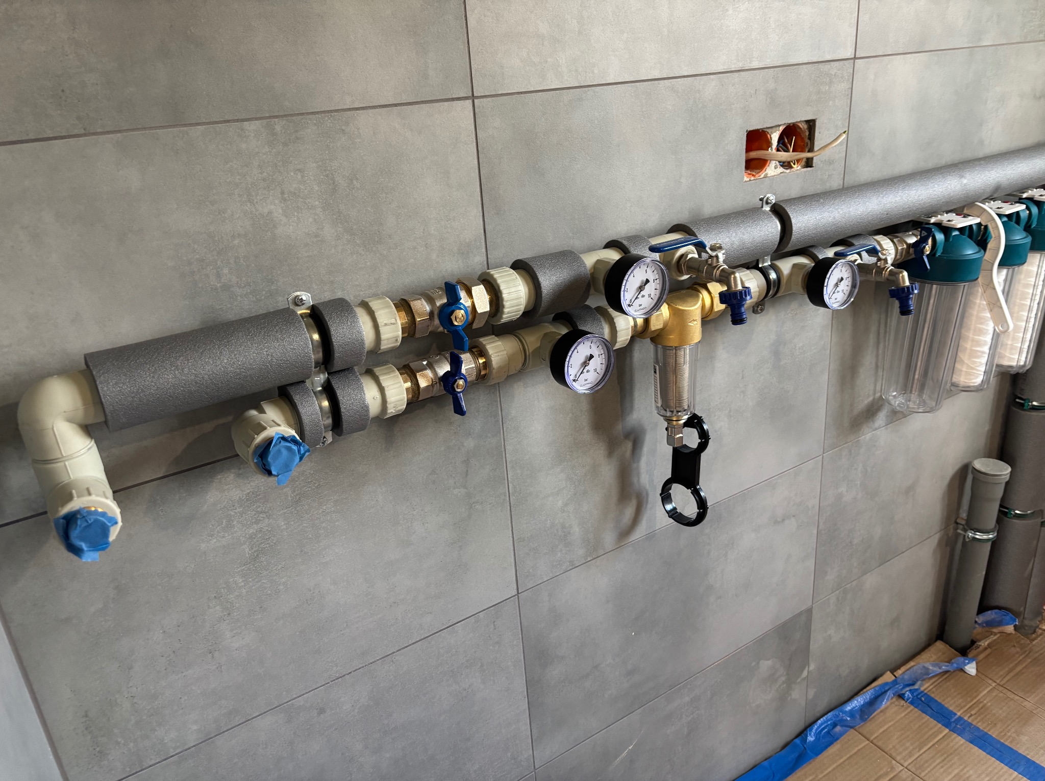 Szereg rur hydraulicznych z izolacją termiczną i manometrami zamontowany na szarej ścianie z płytek, widoczne filtry wody i otwarte gniazdko elektryczne.