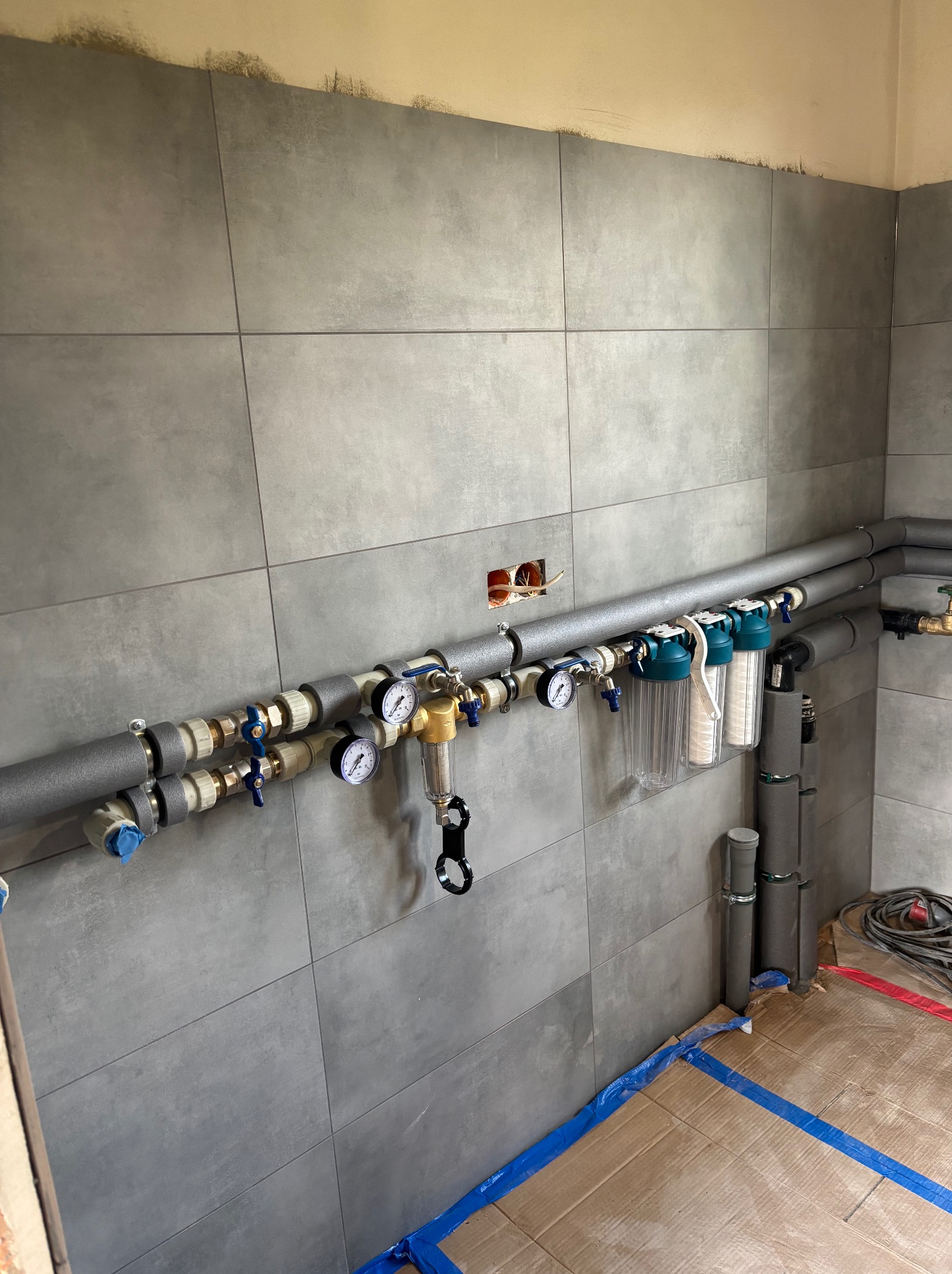 Szare płytki ścienne z widoczną instalacją hydrauliczną: rury z izolacją, zawory, manometry i filtry wody. Podłoga zabezpieczona kartonem i folią.