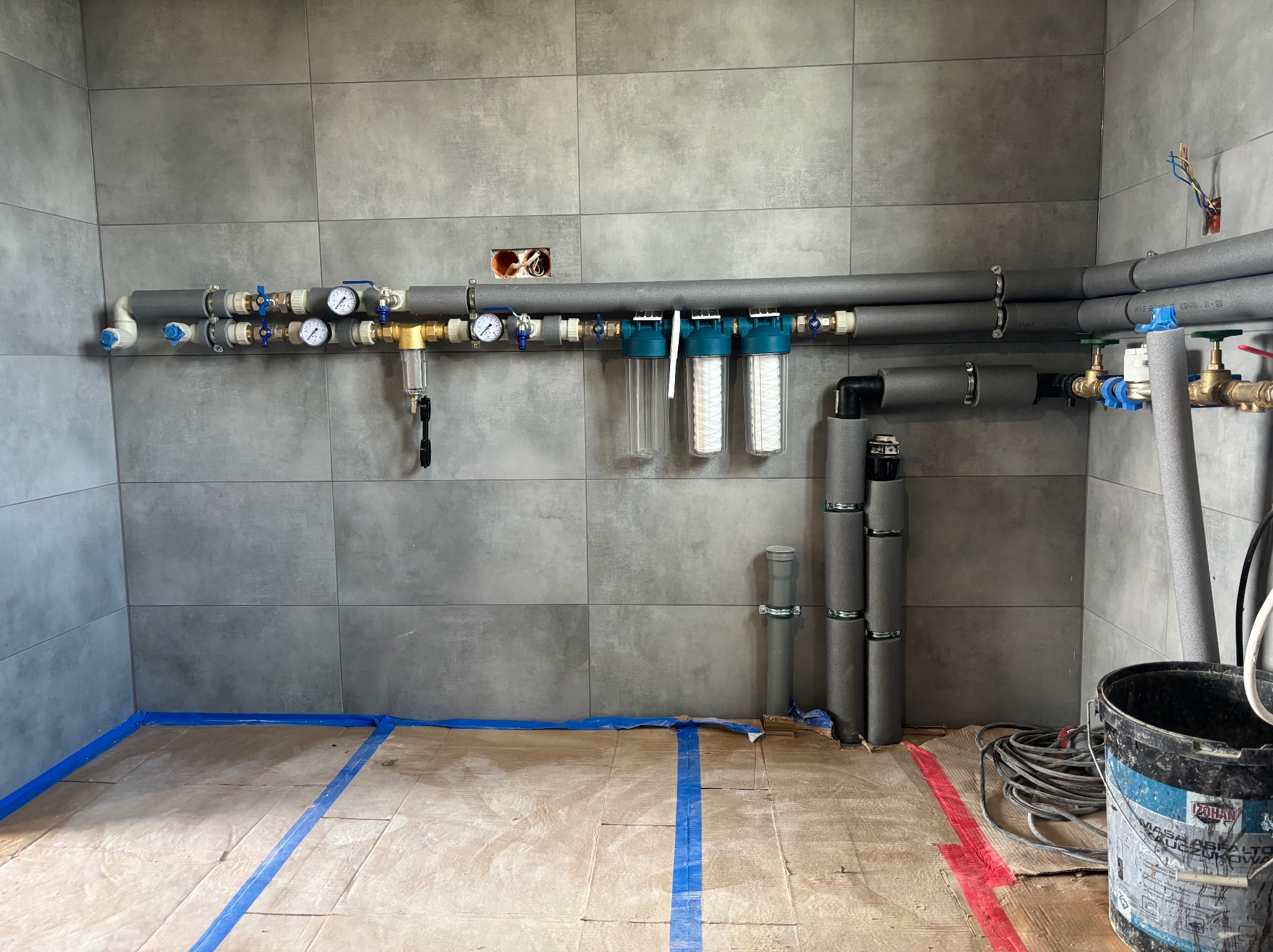 Szare rury instalacji wodnej z manometrami, zaworami i filtrami wody zamontowane na szarej ścianie z płytek. Podłoga zabezpieczona kartonem i niebieską taśmą malarską. Widoczne przewody elektryczne...