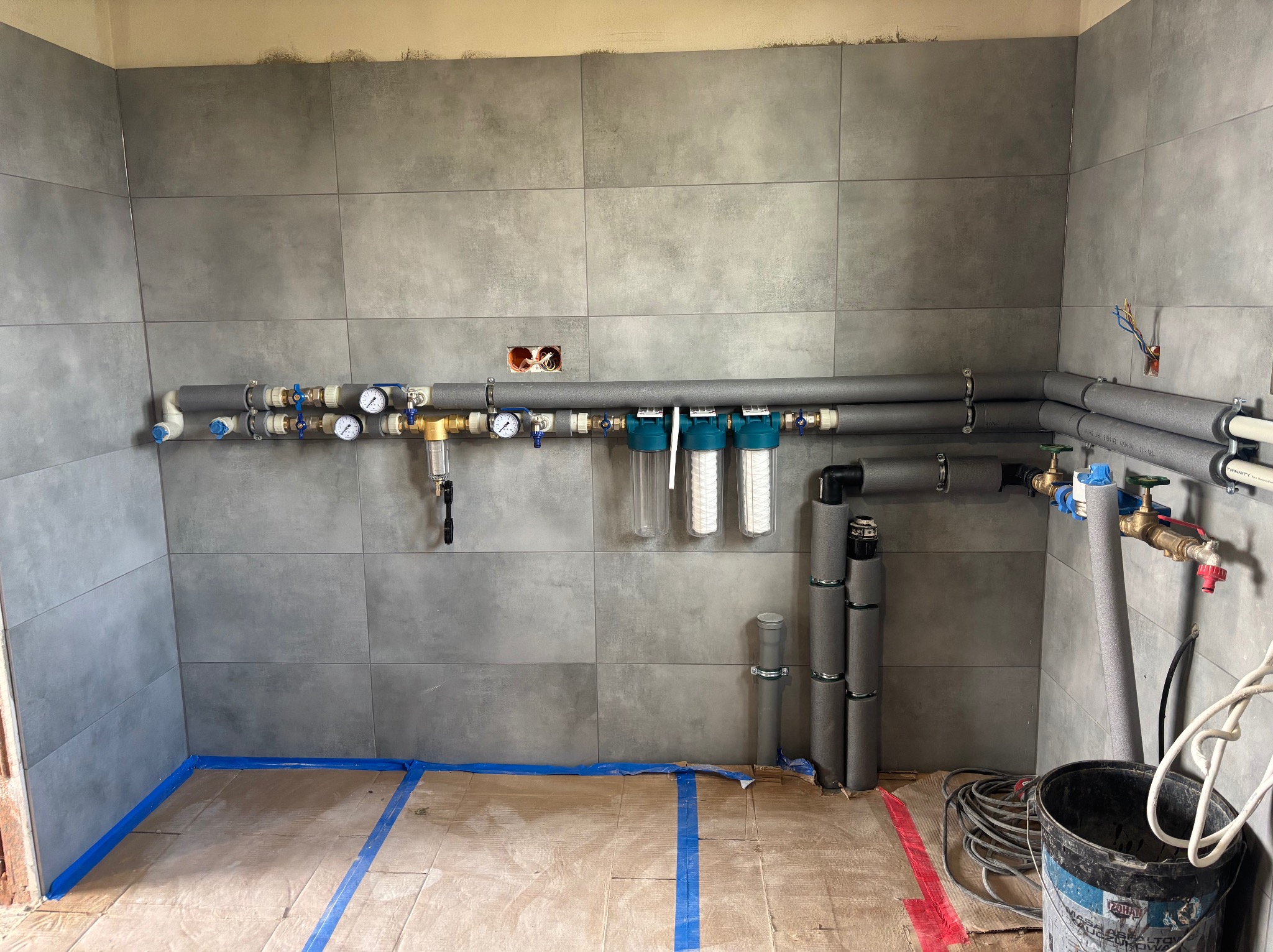 Szare płytki ścienne w trakcie montażu instalacji wodnej z rurami w izolacji, zaworami, manometrami i filtrami. Podłoga zabezpieczona kartonem i niebieską taśmą malarską. Widoczne przewody...