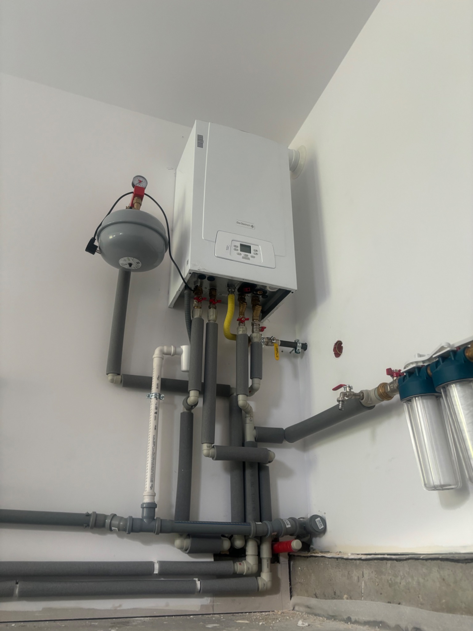 Biały piec gazowy marki De Dietrich zamontowany na ścianie, z widocznymi rurami i zaworami, częściowo zaizolowanymi szarą pianką. Dodatkowo widoczny zbiornik wyrównawczy i filtry wody. Ściany...