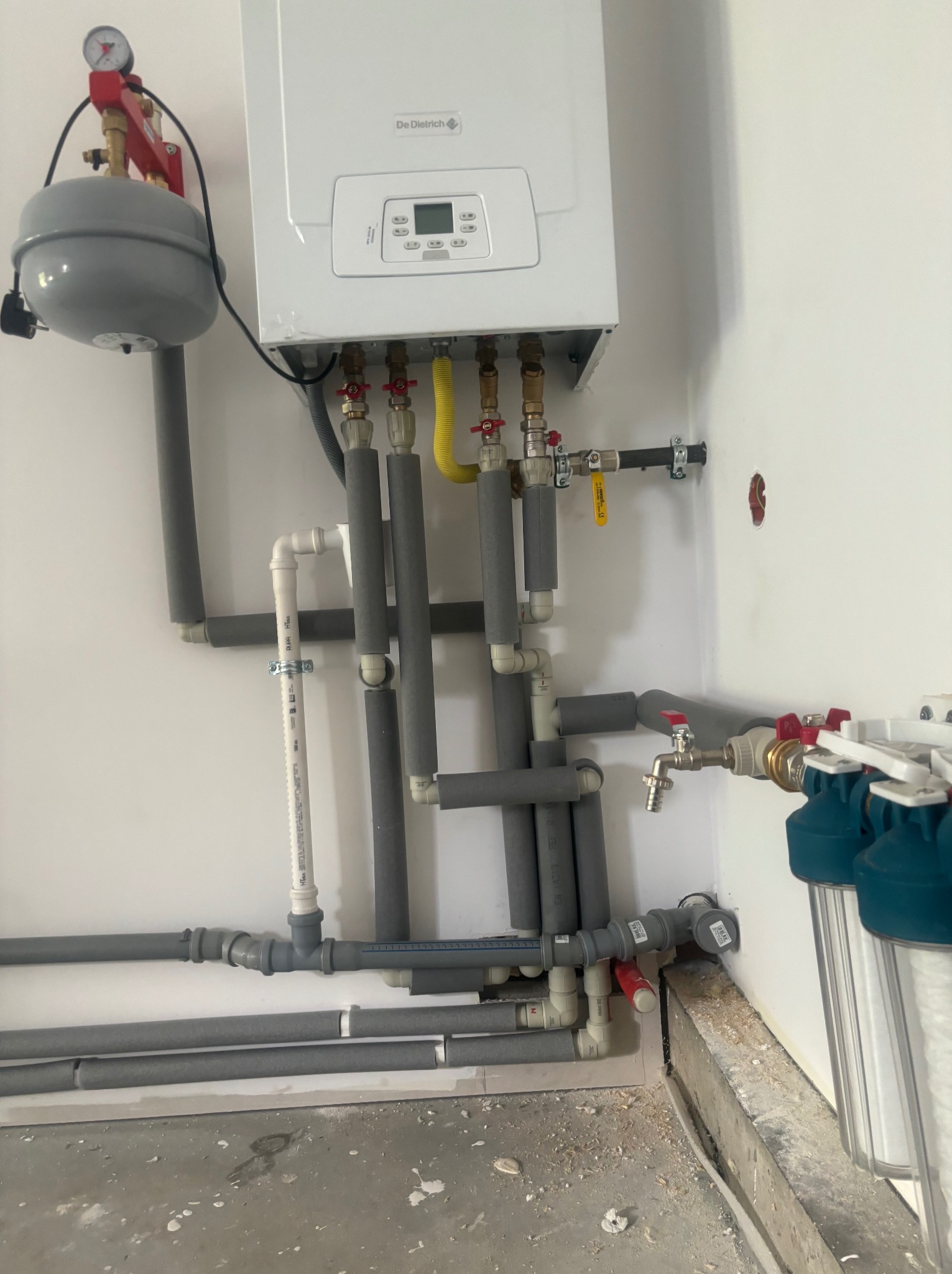 Biały piec gazowy De Dietrich zamontowany na ścianie, z widocznymi rurami, zaworami, manometrem, naczyniem wzbiorczym i filtrami wody, zabezpieczonymi szarą izolacją termiczną.