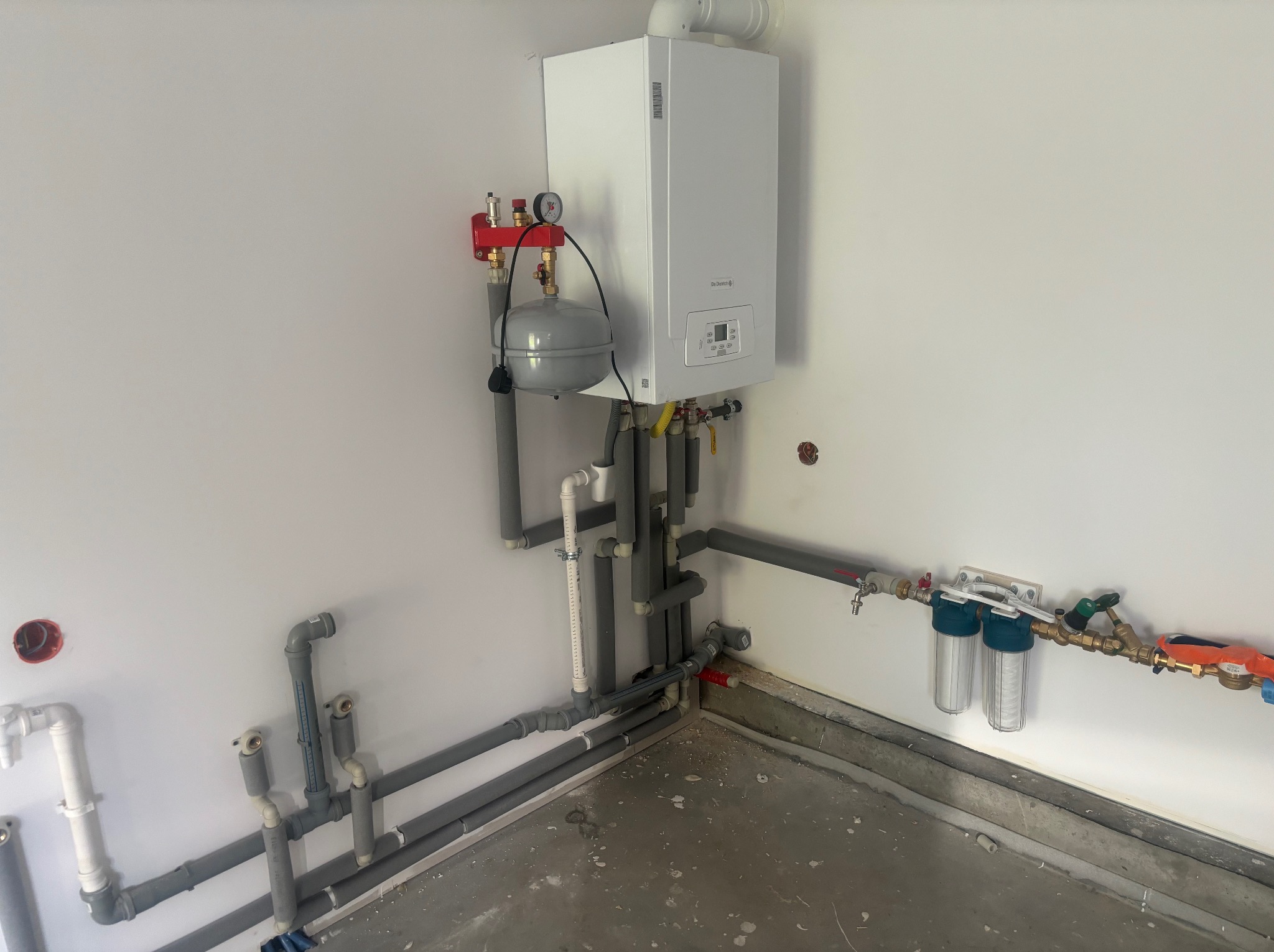 Biały piec gazowy zamontowany na ścianie, z widocznymi rurami, zaworami i filtrami wody. Szare rury rozprowadzone wzdłuż ściany i posadzki. Widoczne elementy instalacji centralnego ogrzewania...