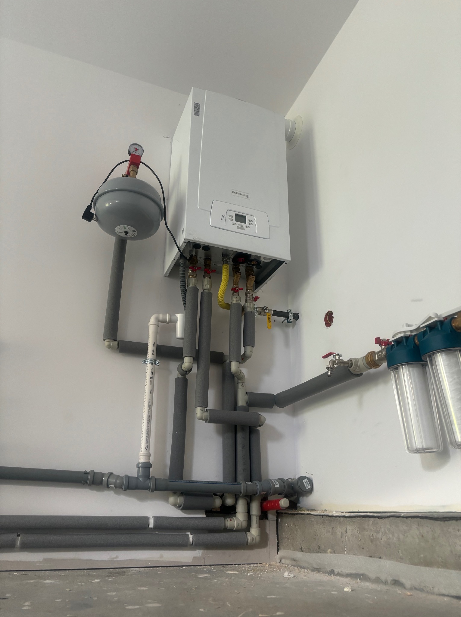 Biały piec gazowy zamontowany na ścianie z szarymi rurami, zaworami i filtrami wody, widok z dołu.
