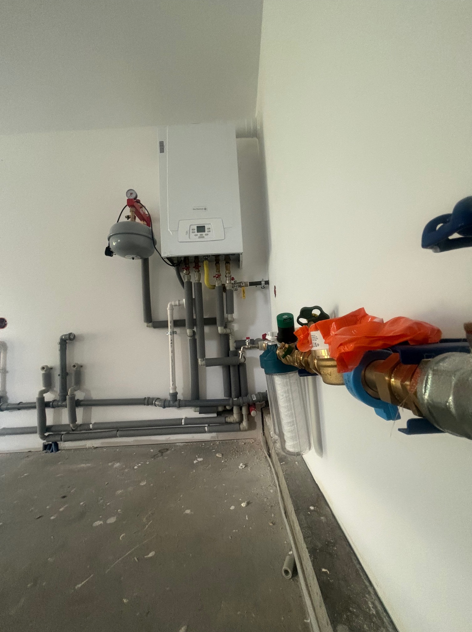 Nowoczesny piec gazowy zamontowany na białej ścianie, z widocznymi rurami i filtrami wody, oraz szarym zbiornikiem wyrównawczym. Podłoga surowa, w trakcie prac wykończeniowych.