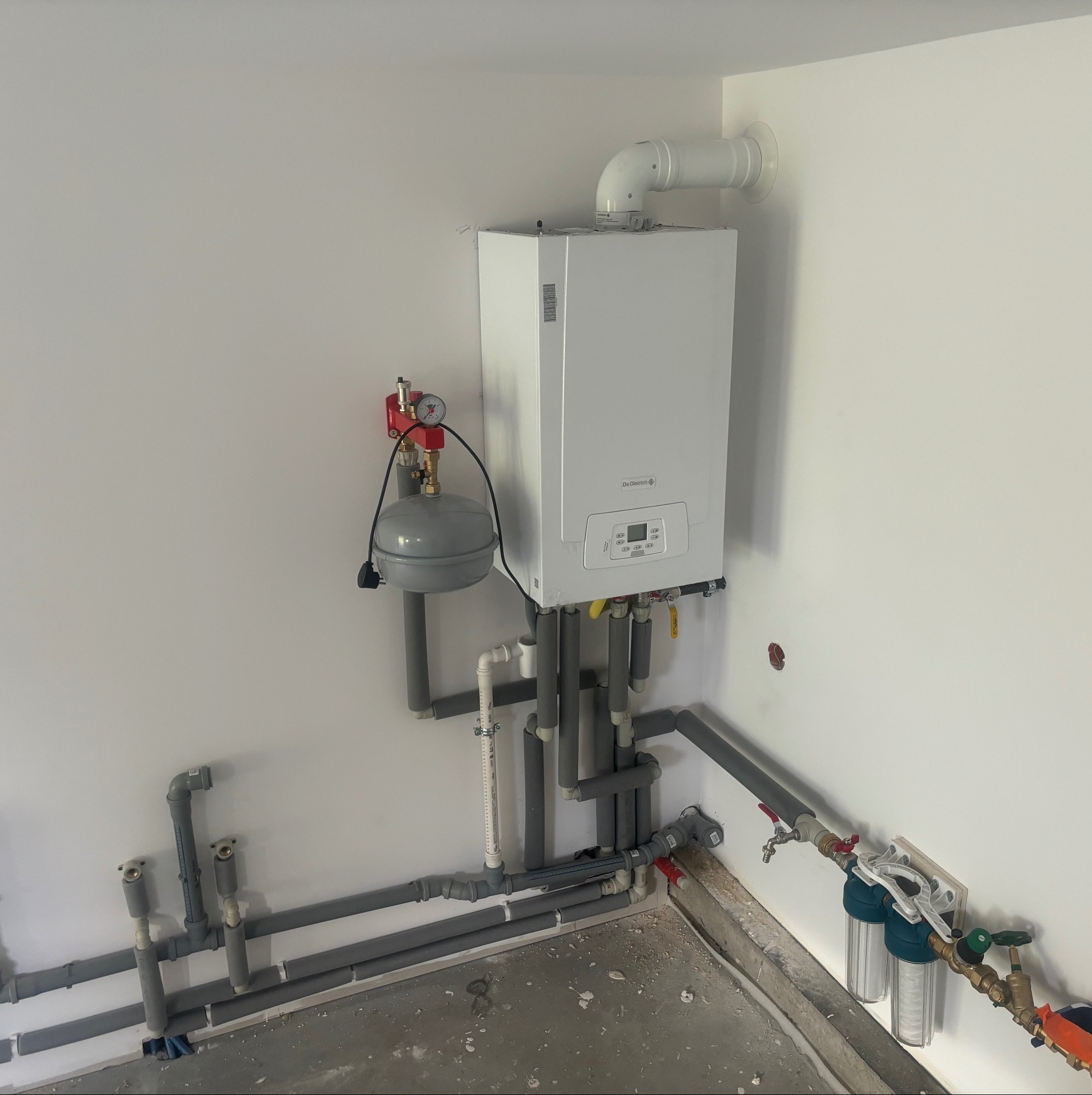 Biały piec gazowy De Dietrich zamontowany na ścianie, z widocznymi szarymi rurami instalacji grzewczej i filtrami wody po prawej stronie, w surowym pomieszczeniu z betonową podłogą.