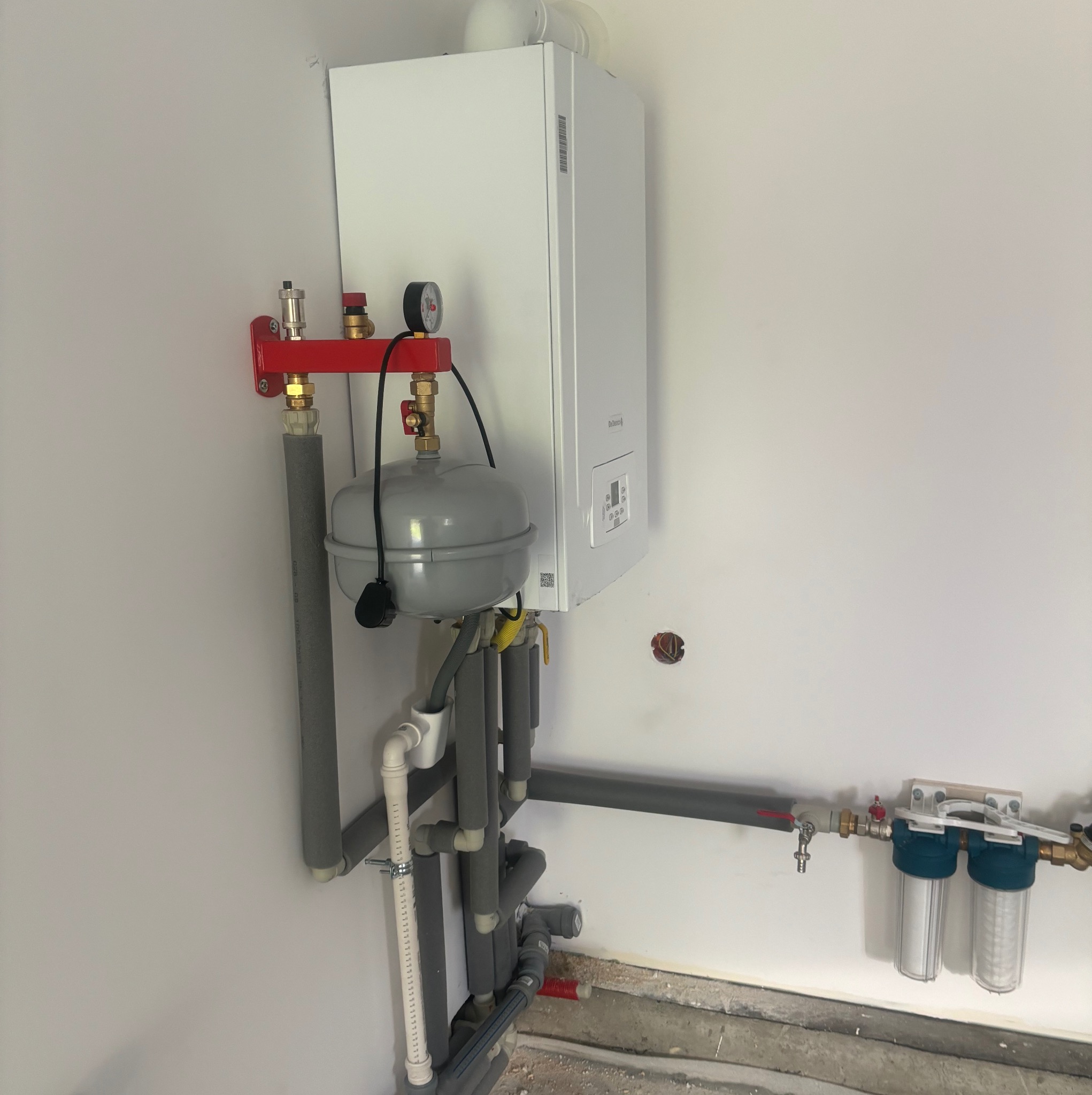 Nowoczesny, biały piec gazowy zamontowany na ścianie z widoczną czerwoną listwą montażową, szarym naczyniem przeponowym i instalacją hydrauliczną z izolowanymi rurami oraz filtrami wody.
