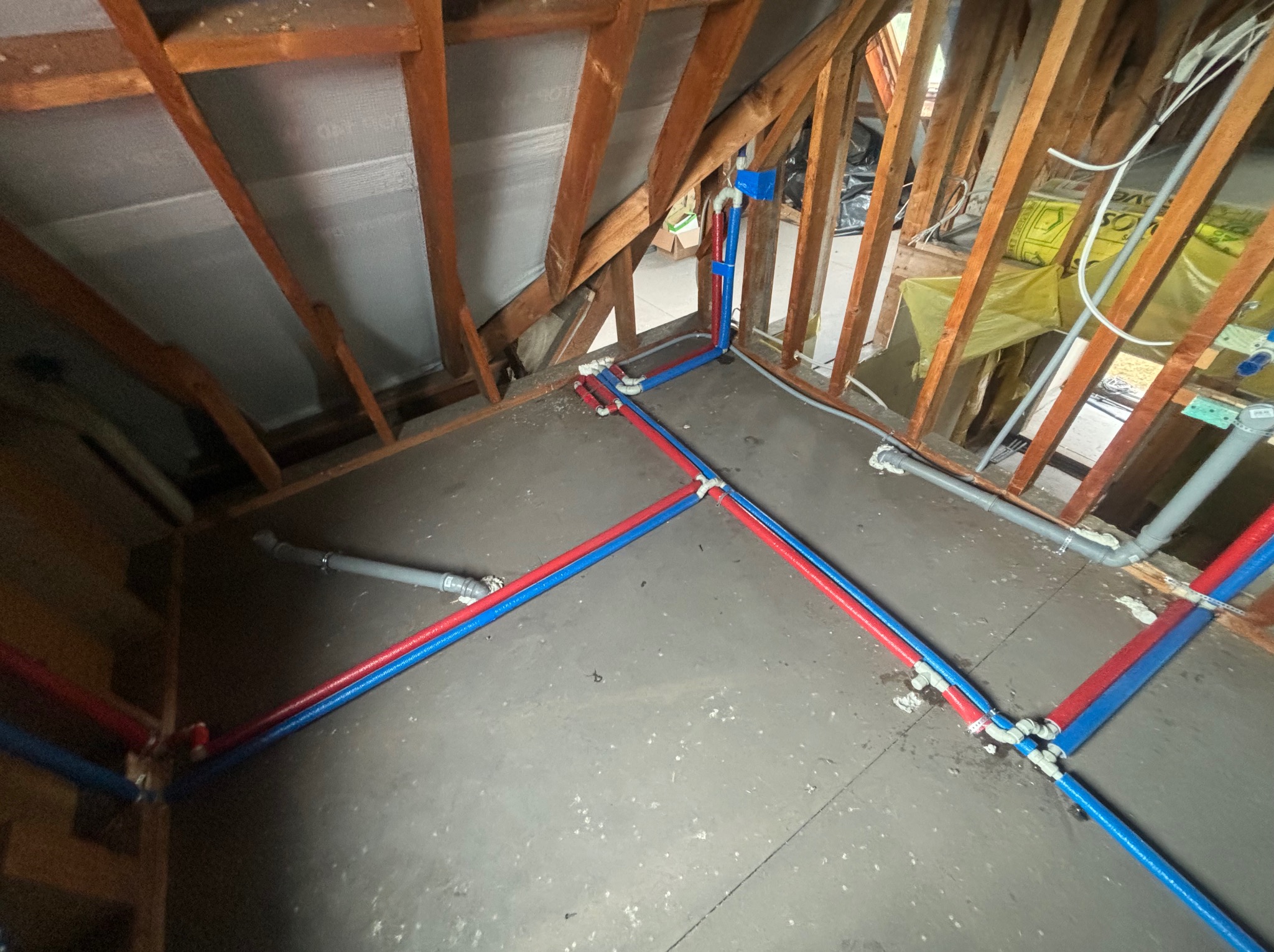 Instalacja wodna z rur PEX w kolorach czerwonym i niebieskim, rozłożona na surowym podłożu w trakcie montażu w przestrzeni poddasza z widoczną konstrukcją drewnianą dachu.