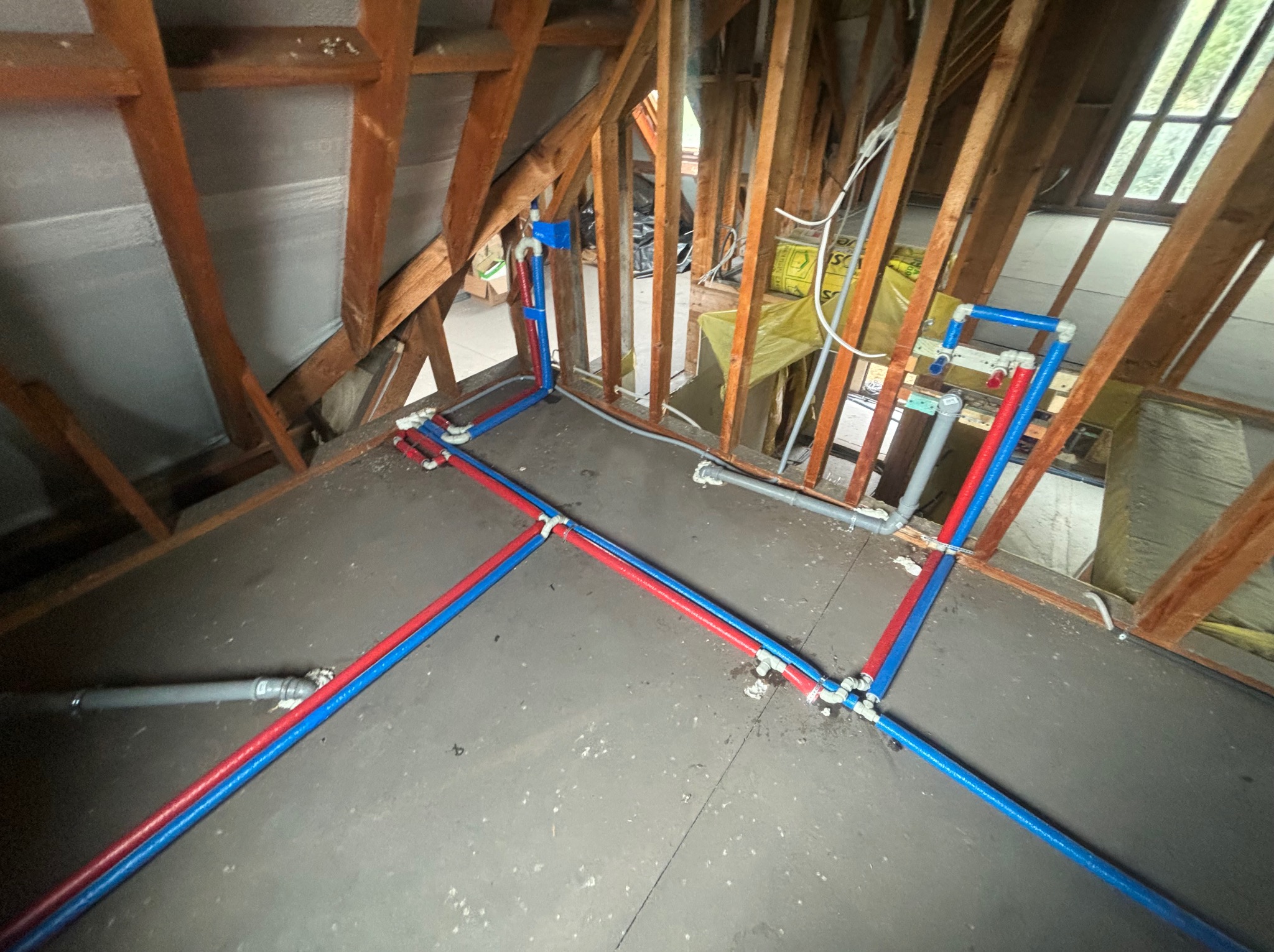 Instalacja wodna z rur PEX w kolorze czerwonym i niebieskim, ułożona w surowym stanie poddasza z widoczną konstrukcją drewnianą, izolacją i oknem w tle.