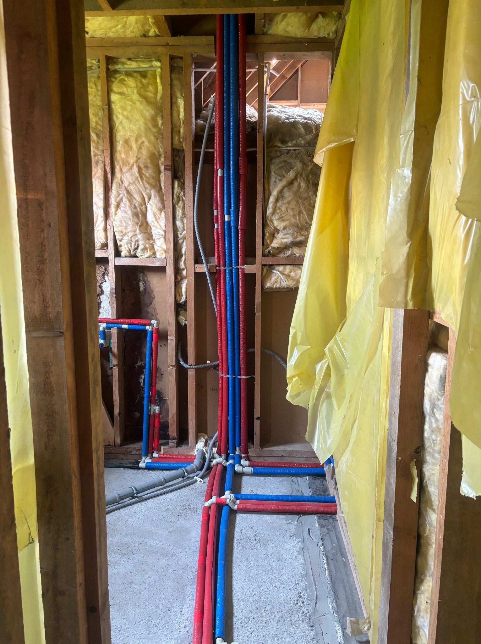 Wnętrze ściany w trakcie montażu instalacji hydraulicznej z widocznymi rurami PEX w kolorach czerwonym i niebieskim, izolacją z wełny mineralnej oraz folią ochronną.