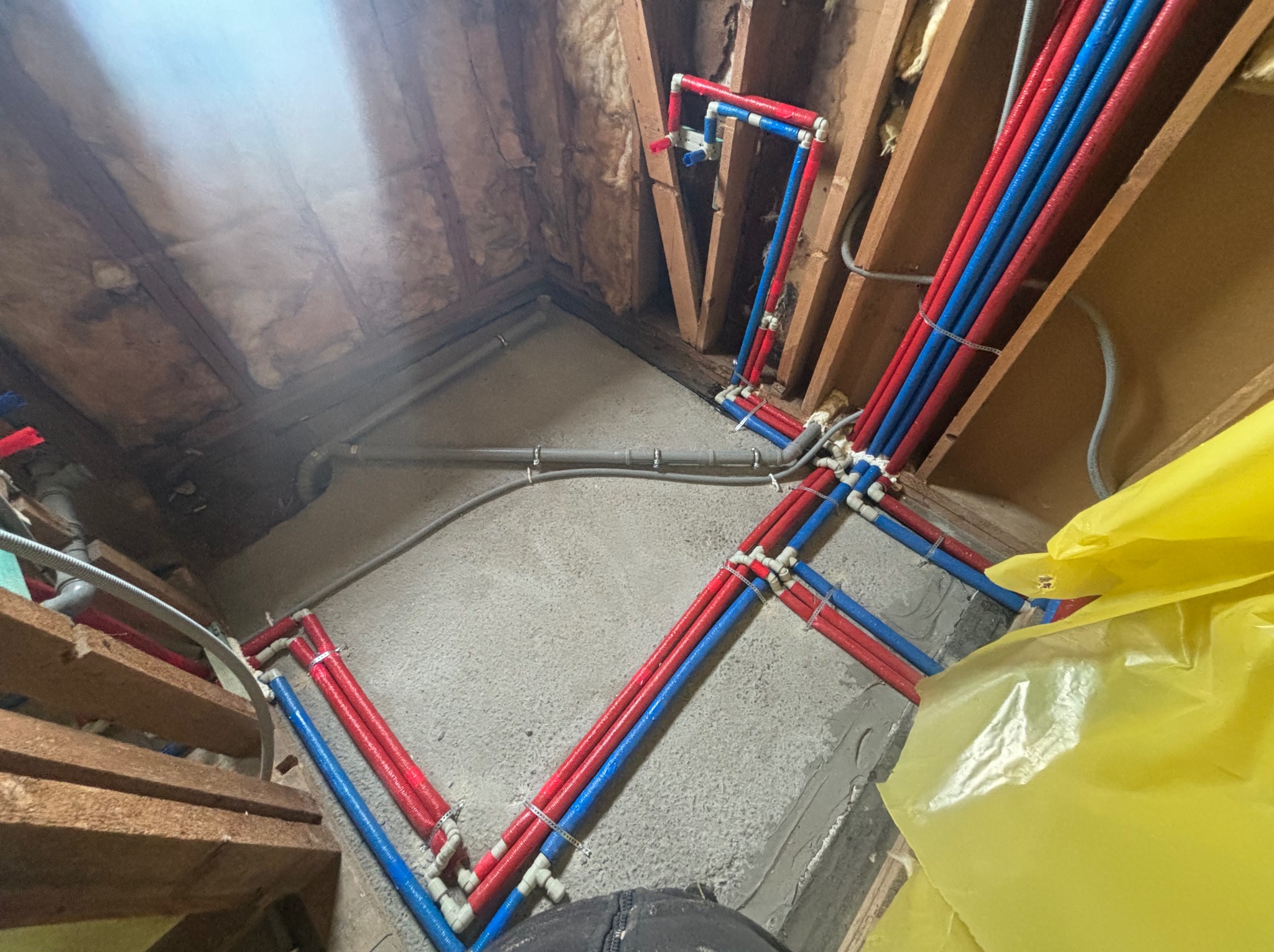 Nowa instalacja wodna z rur PEX w kolorach czerwonym i niebieskim, zamocowana w surowym pomieszczeniu ze ścianami ocieplonymi wełną mineralną, widoczne drewniane elementy konstrukcyjne.