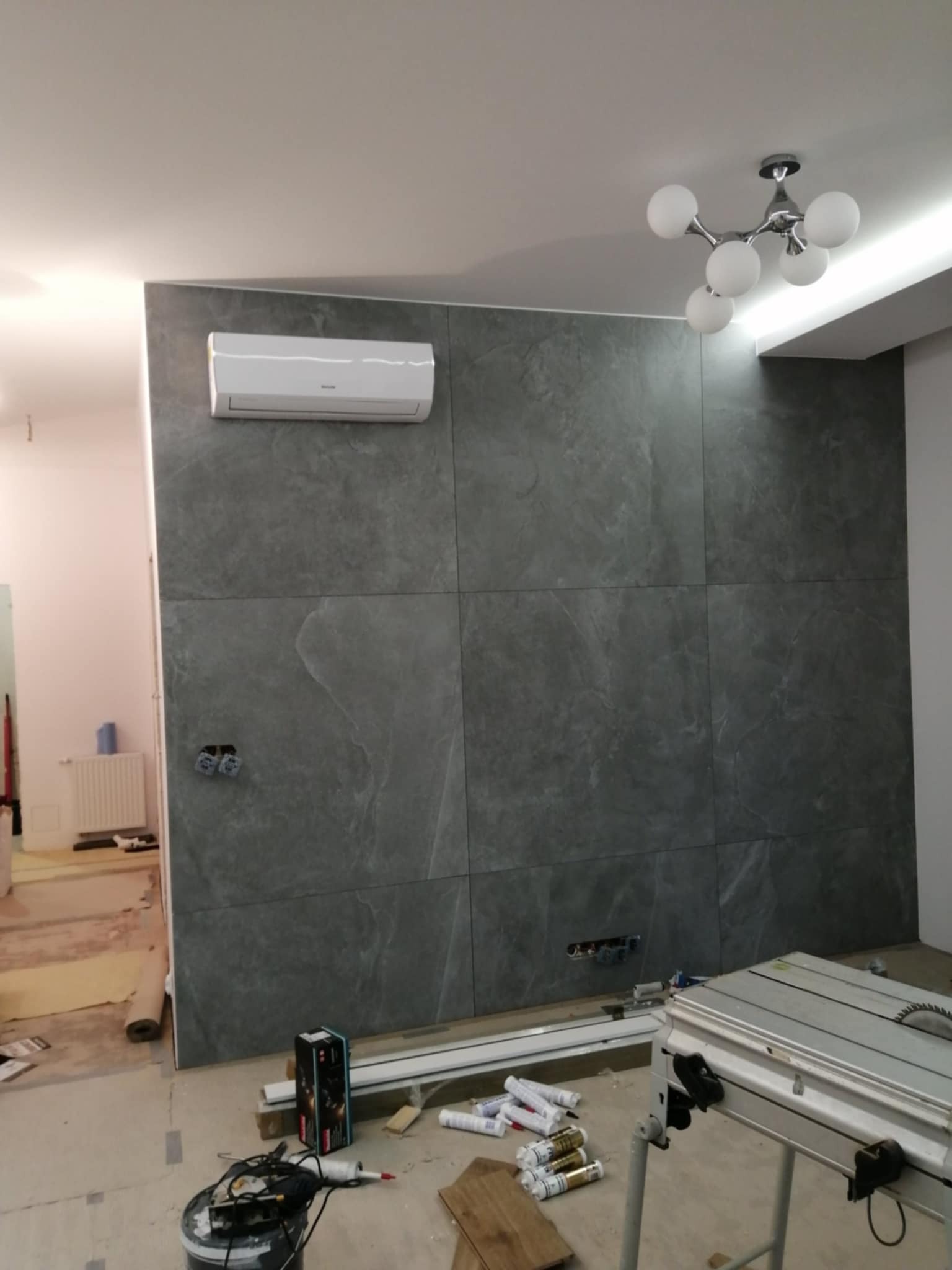 Ściana wyłożona szarą, kwadratową płytką imitującą kamień, z zamontowanym klimatyzatorem. Widoczne gniazdka elektryczne i narzędzia glazurnicze. Nowoczesna lampa sufitowa.