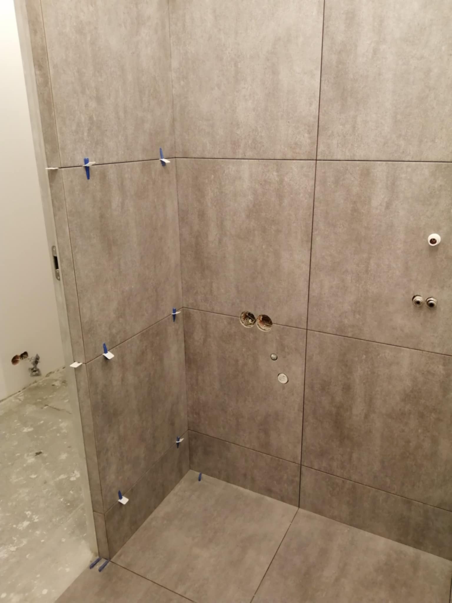 Ściana i podłoga w trakcie układania szarych płytek w łazience. Widoczne krzyżyki dystansowe i otwory na instalacje.