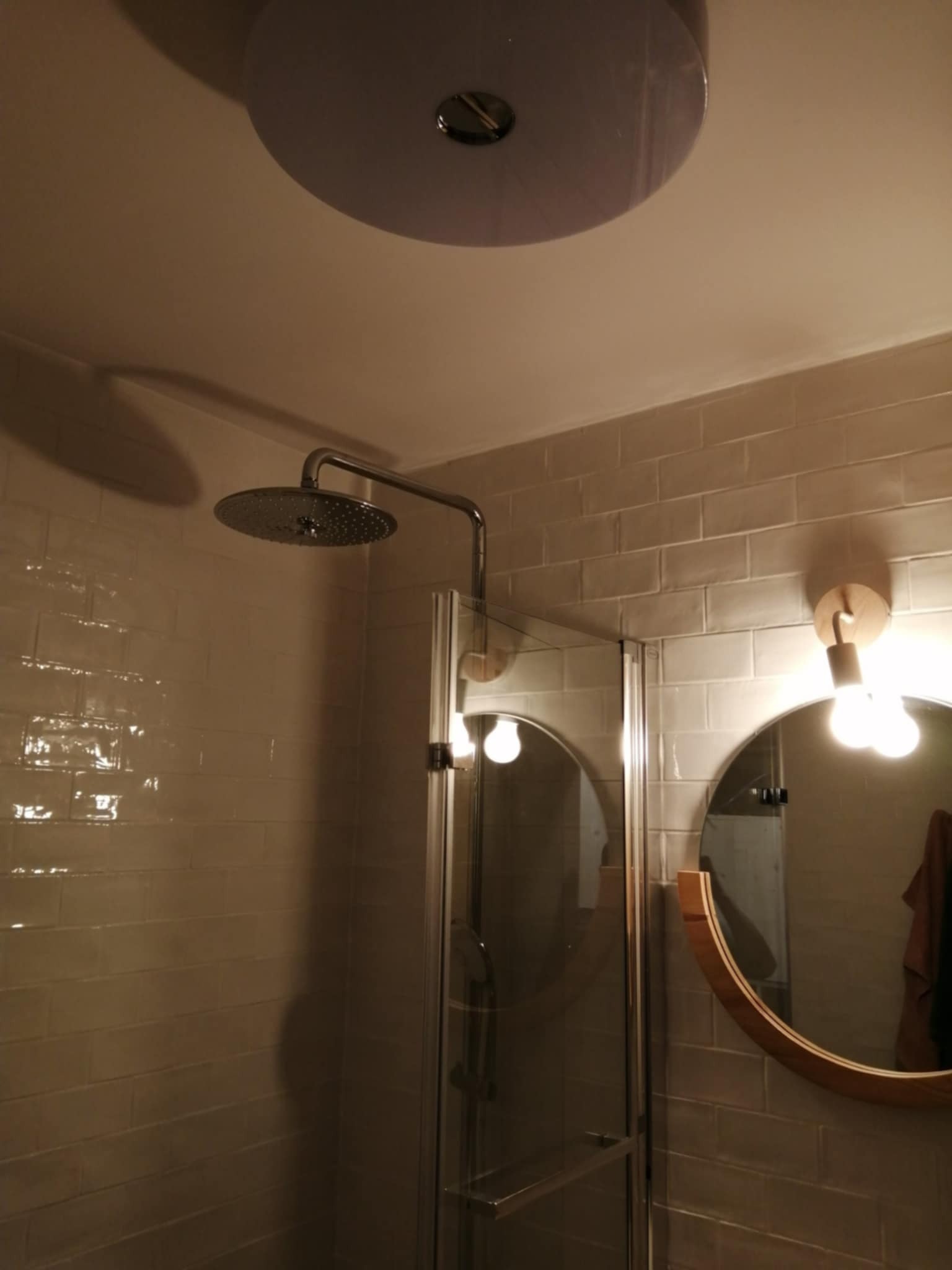 Wykończona kabina prysznicowa z deszczownicą i okrągłym lustrem z drewnianą ramą. Beżowe płytki na ścianach. Widoczne oświetlenie sufitowe.