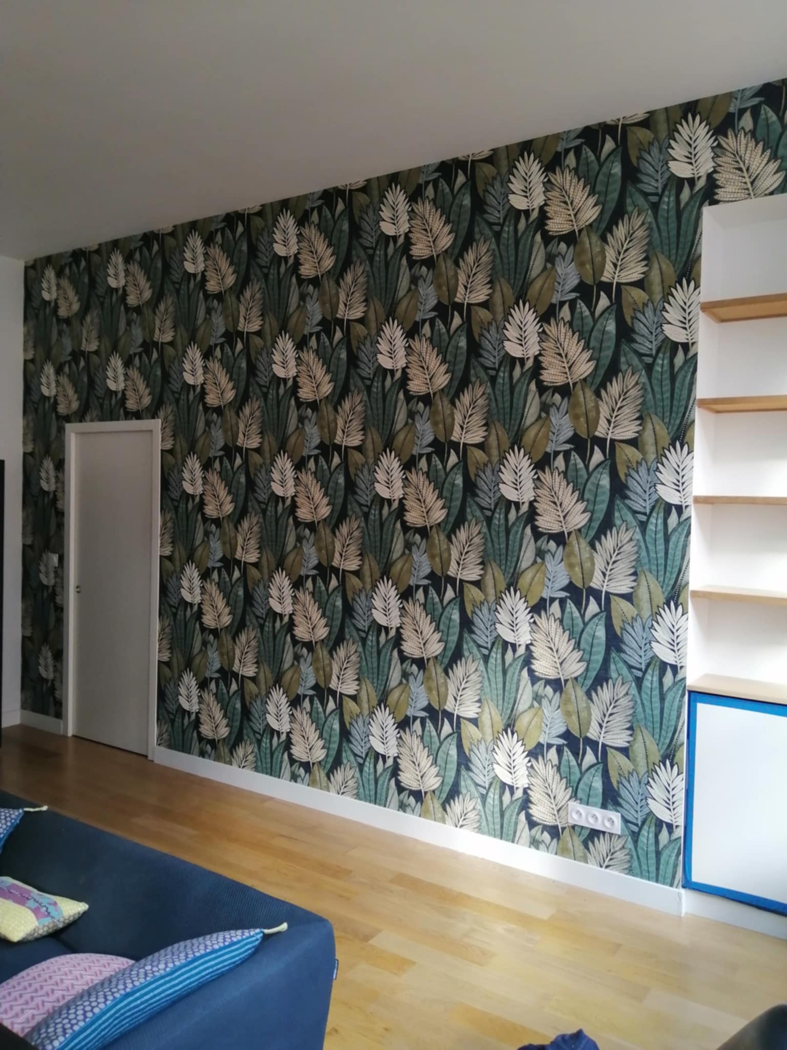 Ściana z tapetą w motywy liściaste, białe drzwi po lewej, regał z półkami po prawej stronie. Jasna podłoga z drewna, widoczny fragment niebieskiej kanapy.