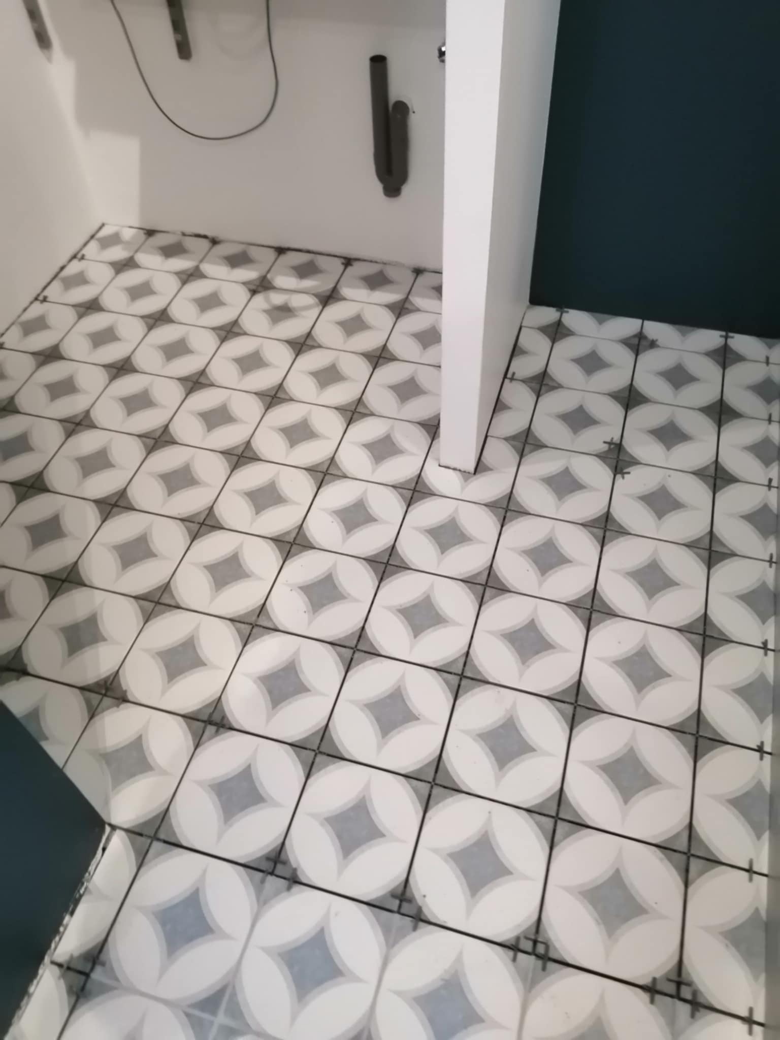 Ułożona podłoga z płytek w geometryczny wzór w łazience, widoczne krzyżyki dystansowe, białe ściany i fragment ciemnej ściany.