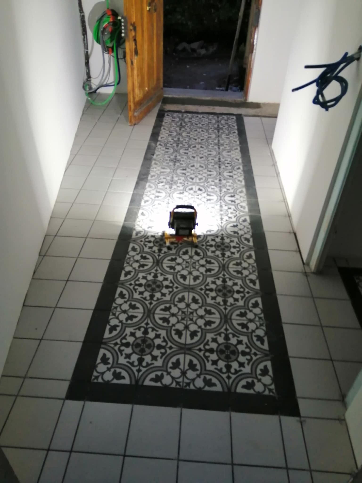 Ułożona podłoga z płytek w korytarzu, wzór geometryczny. Na środku lampa budowlana, oświetlająca fragment posadzki. Widoczne kable i otwarta drewniana rama drzwi.