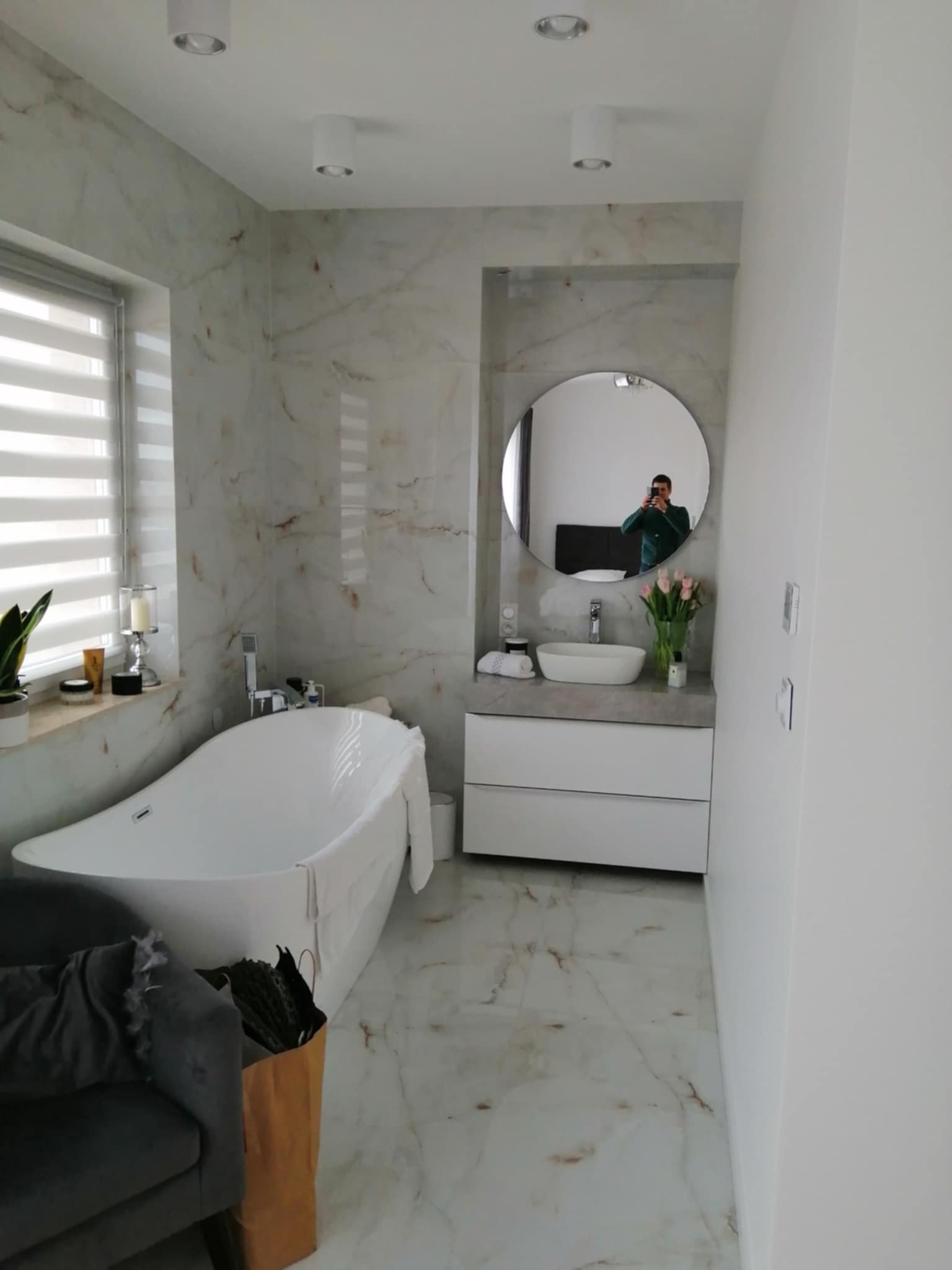 Elegancka łazienka z wanną wolnostojącą, marmurowymi płytkami i okrągłym lustrem, w którym odbija się osoba robiąca zdjęcie. Nowoczesne oświetlenie punktowe.