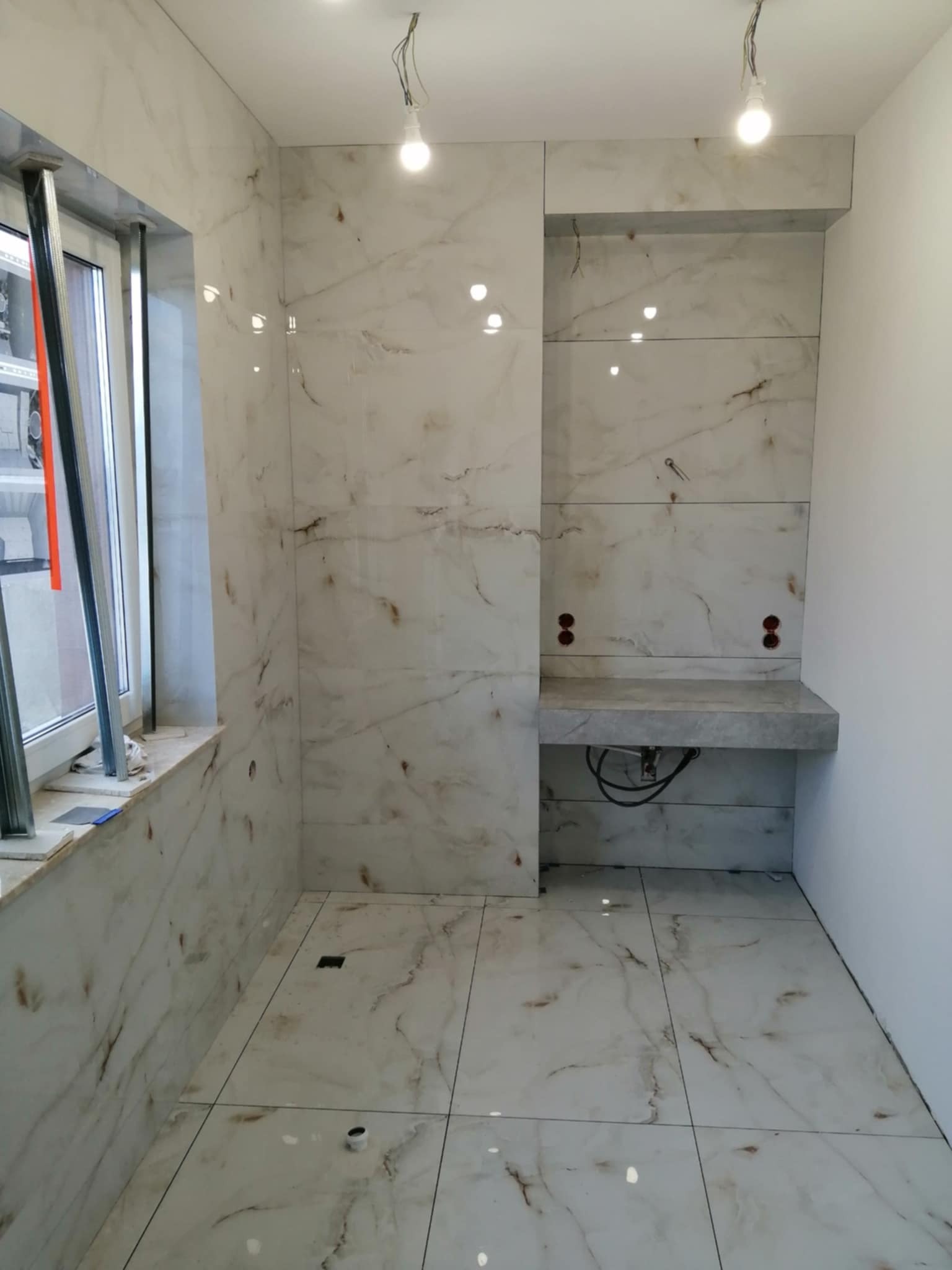 Wykończona łazienka z marmurowymi płytkami. Widoczny fragment okna, zabudowa pod umywalkę i oświetlenie. Płytki ułożone na podłodze i ścianach.