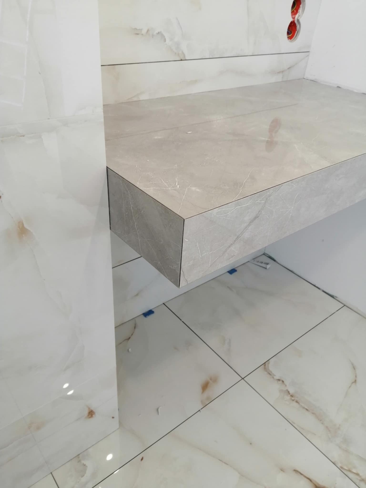 Wykończona, beżowa półka z płytek w łazience, z widocznymi detalami fug i wzoru marmuru. Płytki podłogowe w tym samym stylu, fragment ściany z gniazdami elektrycznymi.