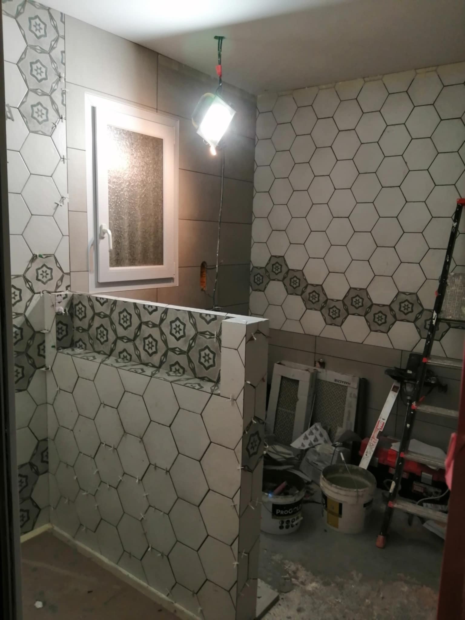 Układanie heksagonalnych płytek w łazience, widoczne narzędzia i drabina. Częściowo ułożone płytki z wzorem kwiatowym. Praca w toku, oświetlenie halogenowe.
