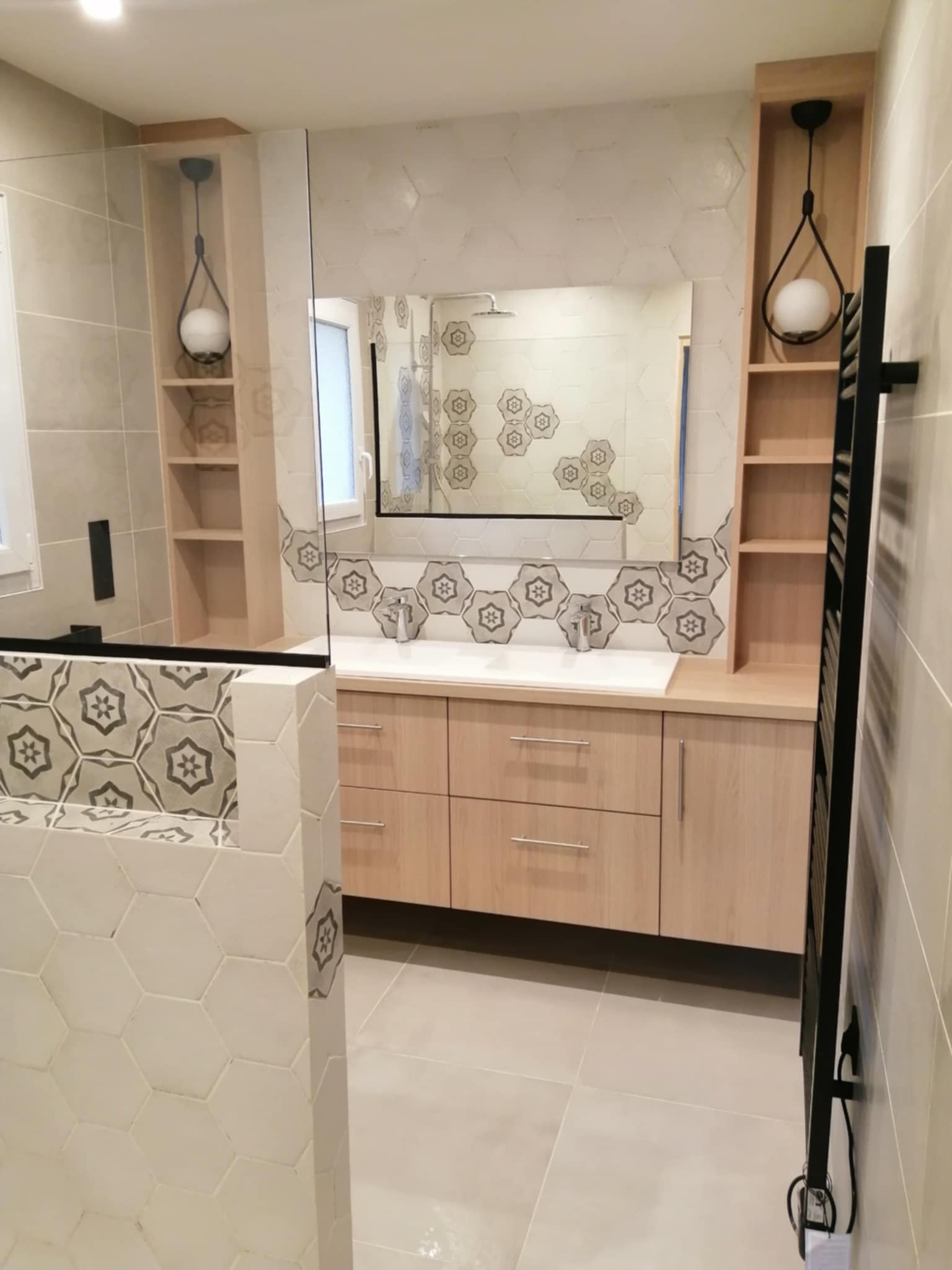 Jasna łazienka po remoncie z heksagonalnymi płytkami, drewnianą szafką pod umywalką, lustrem i czarnym grzejnikiem drabinkowym. Nowoczesne oświetlenie.