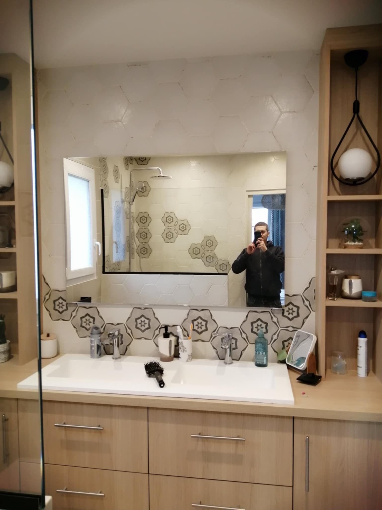 Ukończona łazienka z umywalką, szafkami z jasnego drewna i heksagonalnymi płytkami. W lustrze odbija się osoba robiąca zdjęcie. Lampy wiszące i półki dodają stylu.