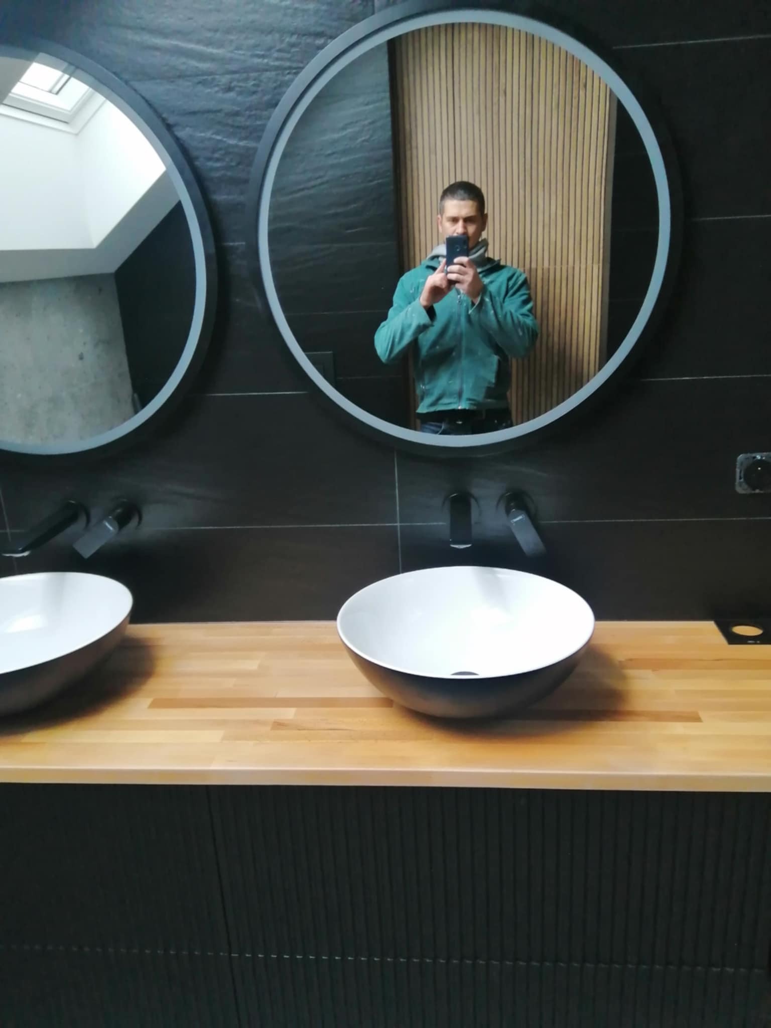 Nowoczesna łazienka z dwoma okrągłymi lustrami, drewnianym blatem i czarnymi umywalkami. W lustrze odbija się osoba robiąca zdjęcie.