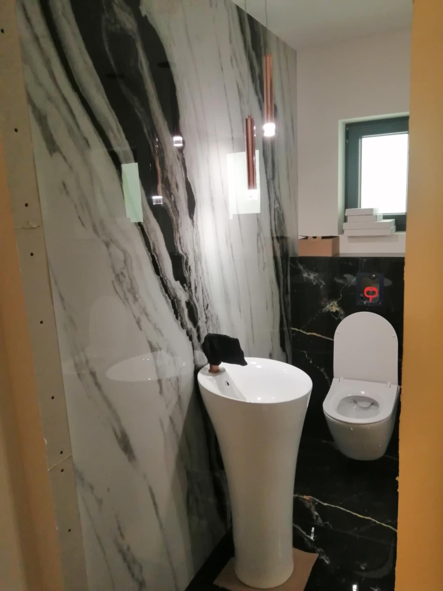 Wnętrze łazienki w trakcie wykańczania: marmurowe płytki na ścianach, biała umywalka na postumencie i toaleta. Widoczne elementy instalacji i oświetlenie.