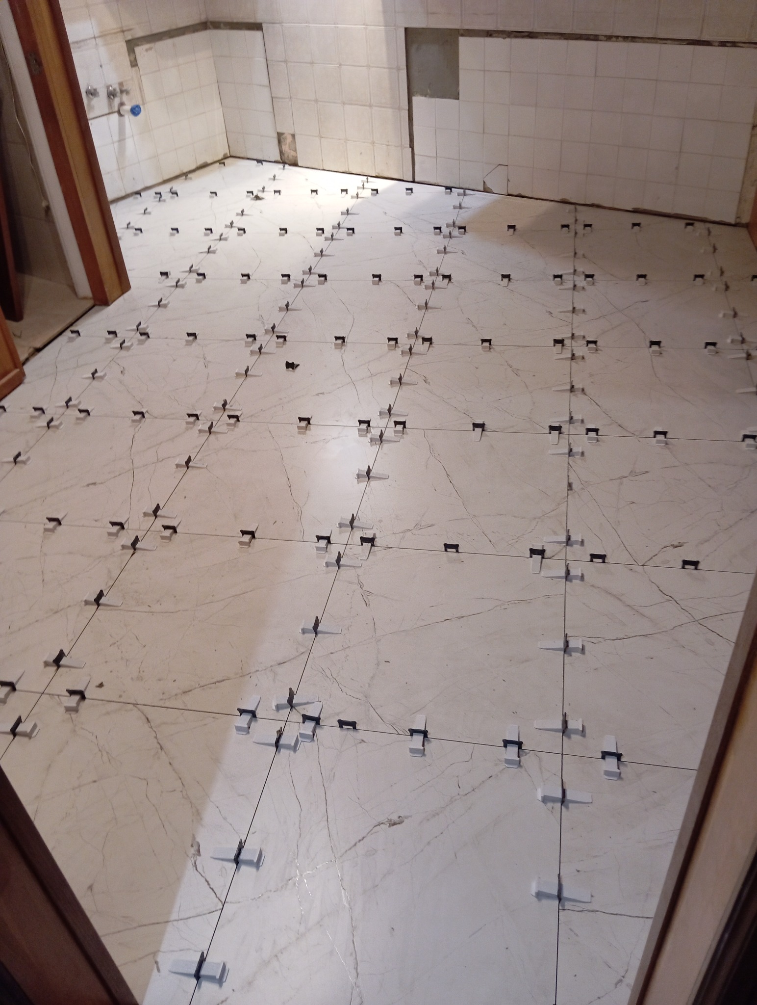 Układanie płytek podłogowych w łazience z użyciem systemu poziomowania płytek. Widoczne krzyżyki dystansowe i płytki imitujące marmur.
