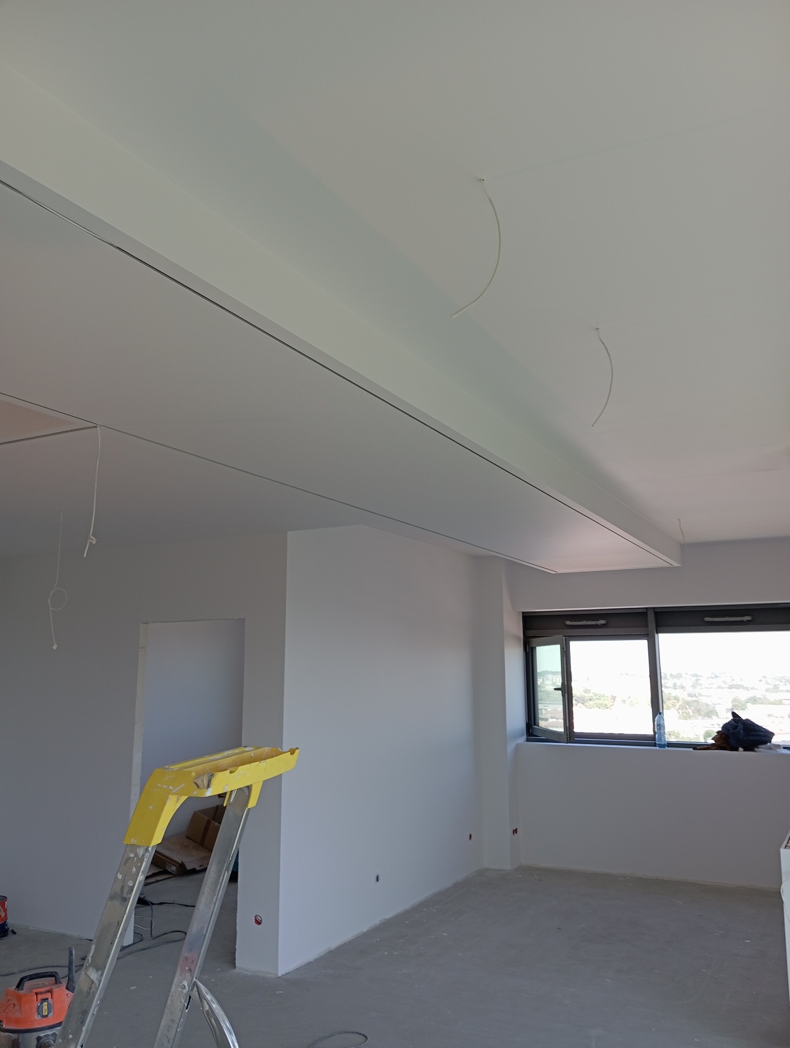 Białe wnętrze w trakcie prac wykończeniowych: ściany pomalowane na biało, widoczna drabina, okno z widokiem na miasto. Sufit z zamontowanym oświetleniem.