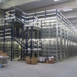 Logis - Wielopoziomowy system regałów magazynowych z metalową konstrukcją, schodami i pomostami, częściowo wypełniony kartonami, widok z perspektywy podłogi magazynu.