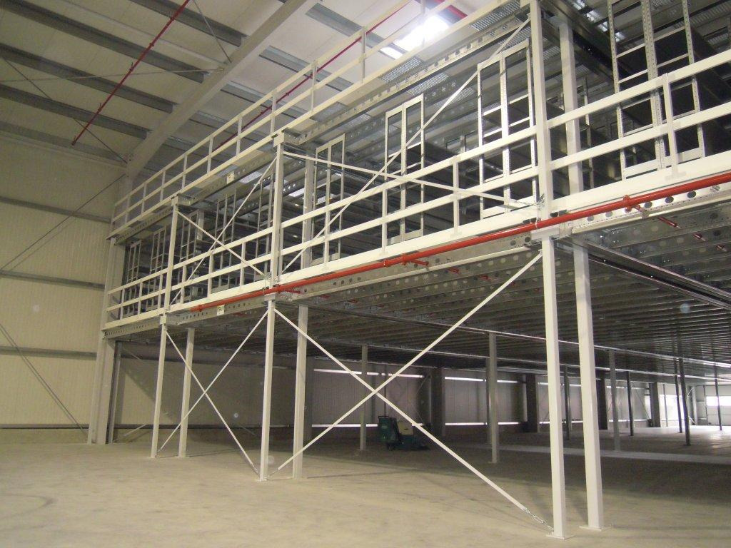 Wnętrze hali magazynowej z antresolą stalową, widoczne białe słupy nośne, czerwona instalacja przeciwpożarowa pod sufitem i betonowa posadzka.