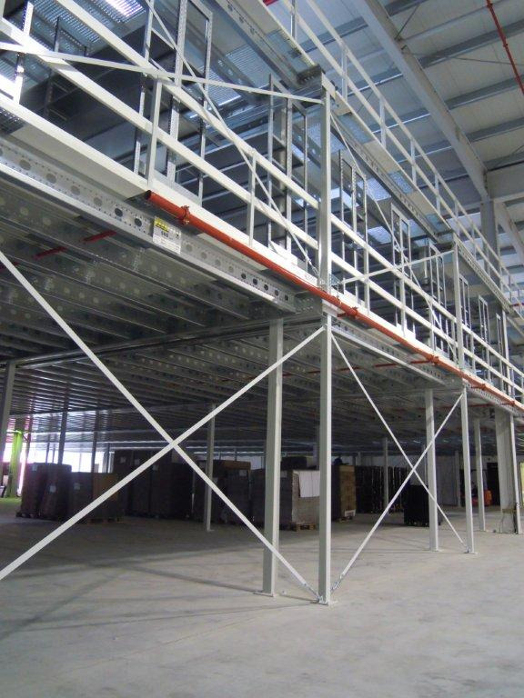 Wnętrze hali magazynowej z widoczną metalową konstrukcją antresoli z pomarańczową rurą, wspartą na białych słupach, z kartonami składowanymi pod spodem.