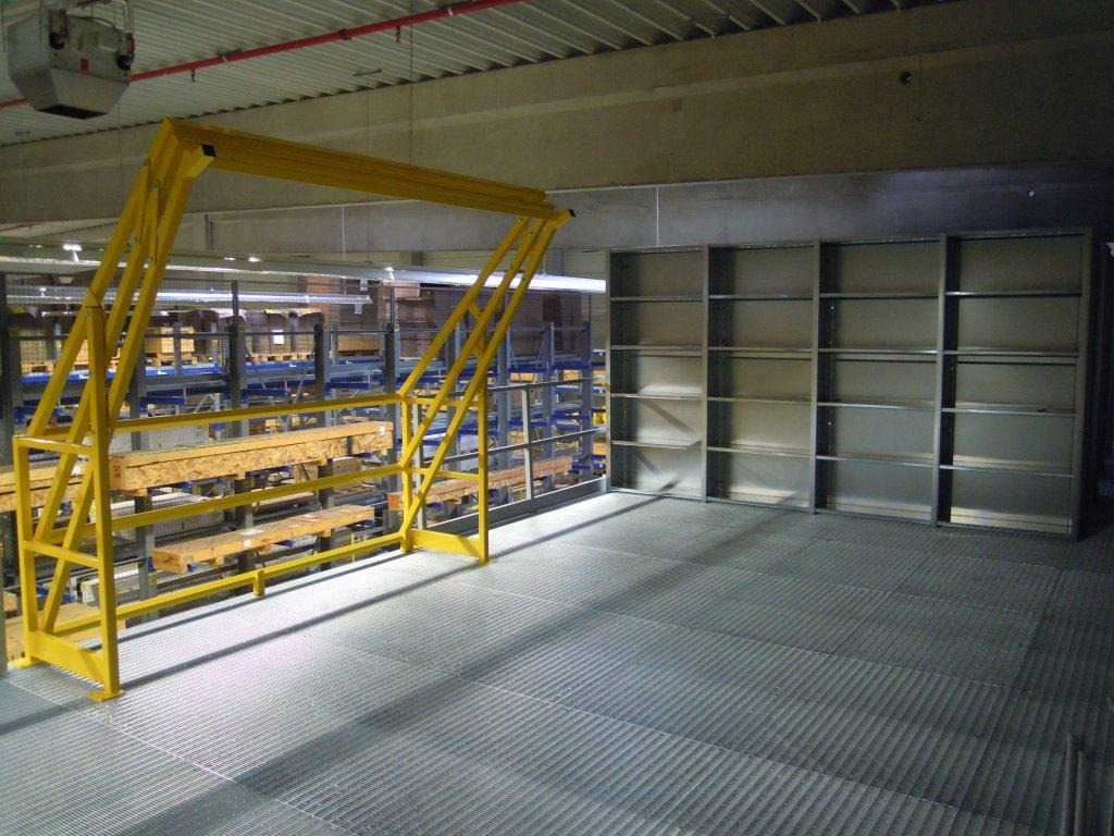 Metalowe regały magazynowe z żółtą konstrukcją zabezpieczającą na antresoli z kratownicową podłogą w hali magazynowej.