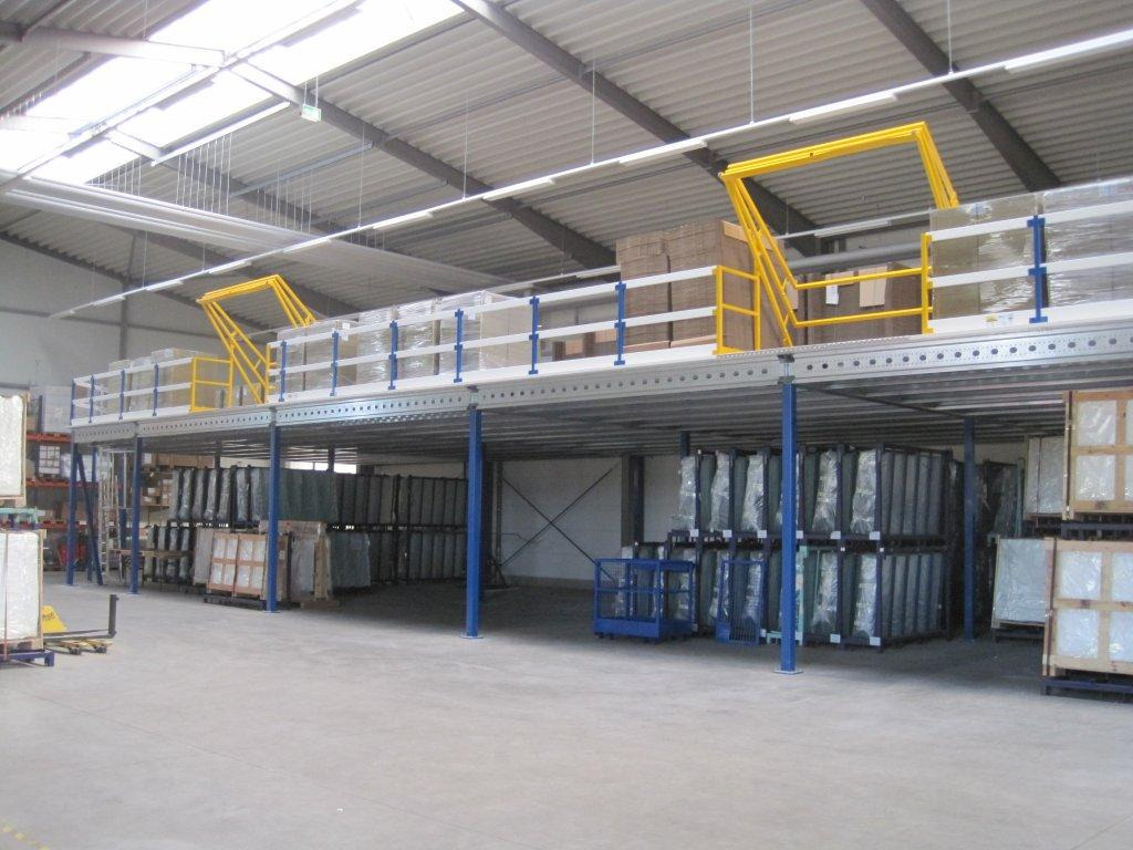 Wnętrze magazynu z antresolą na metalowych słupach, zabezpieczoną niebieskimi barierkami i żółtymi schodami, widoczne towary na paletach i w opakowaniach.