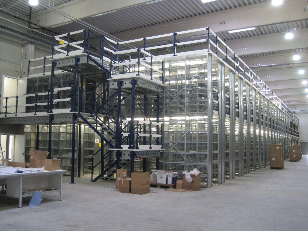 Wielopoziomowy system regałów magazynowych z metalową konstrukcją, schodami i pomostami, częściowo wypełniony kartonami, widok z perspektywy podłogi magazynu.