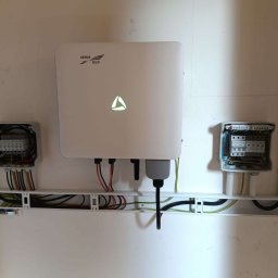 NEXT GENERATION ENERGY SPÓŁKA Z OGRANICZONĄ ODPOWIEDZIALNOŚCIĄ - Biała obudowa falownika z logo Kehua Tech, zamontowana na ścianie z widocznymi kablami i dwiema otwartymi puszkami elektrycznymi po bokach.